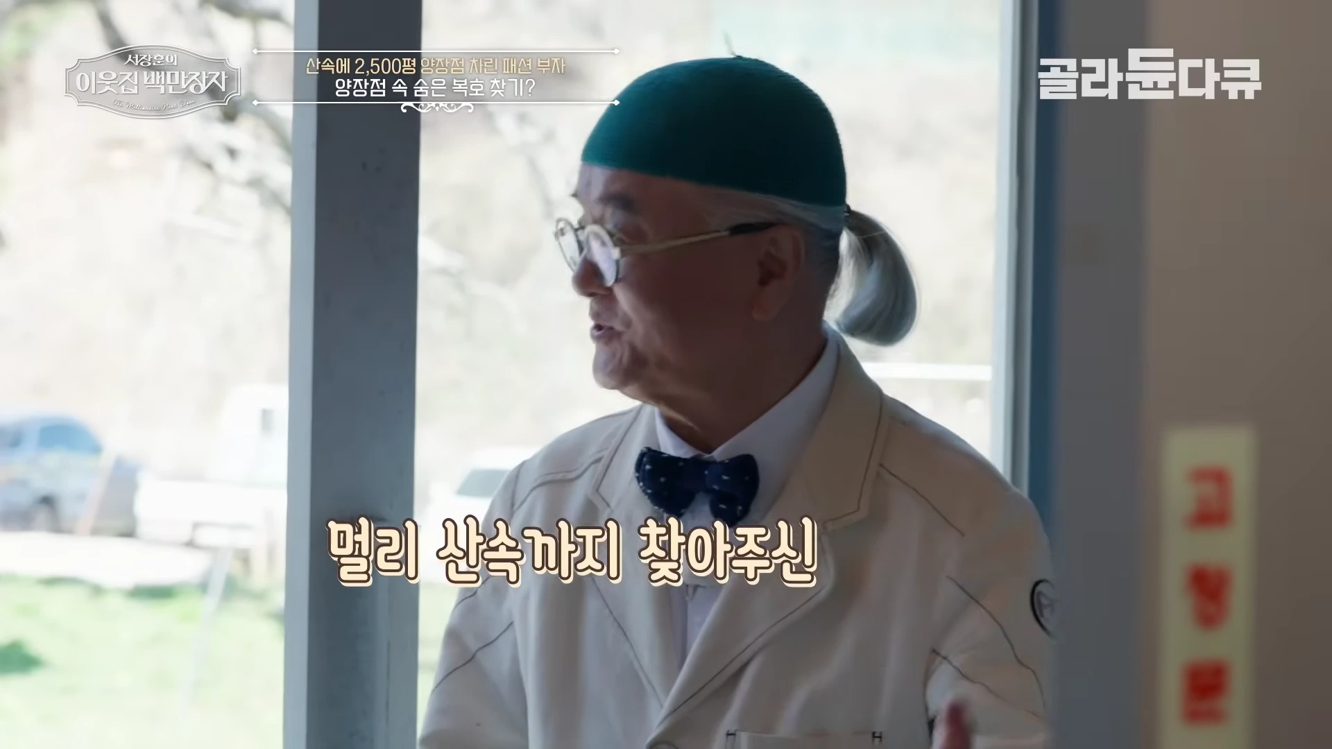 초록색 모자를 쓰고 나비넥타이를 맨 백발의 남성이 실내에서 이야기를 나누고 있는 모습