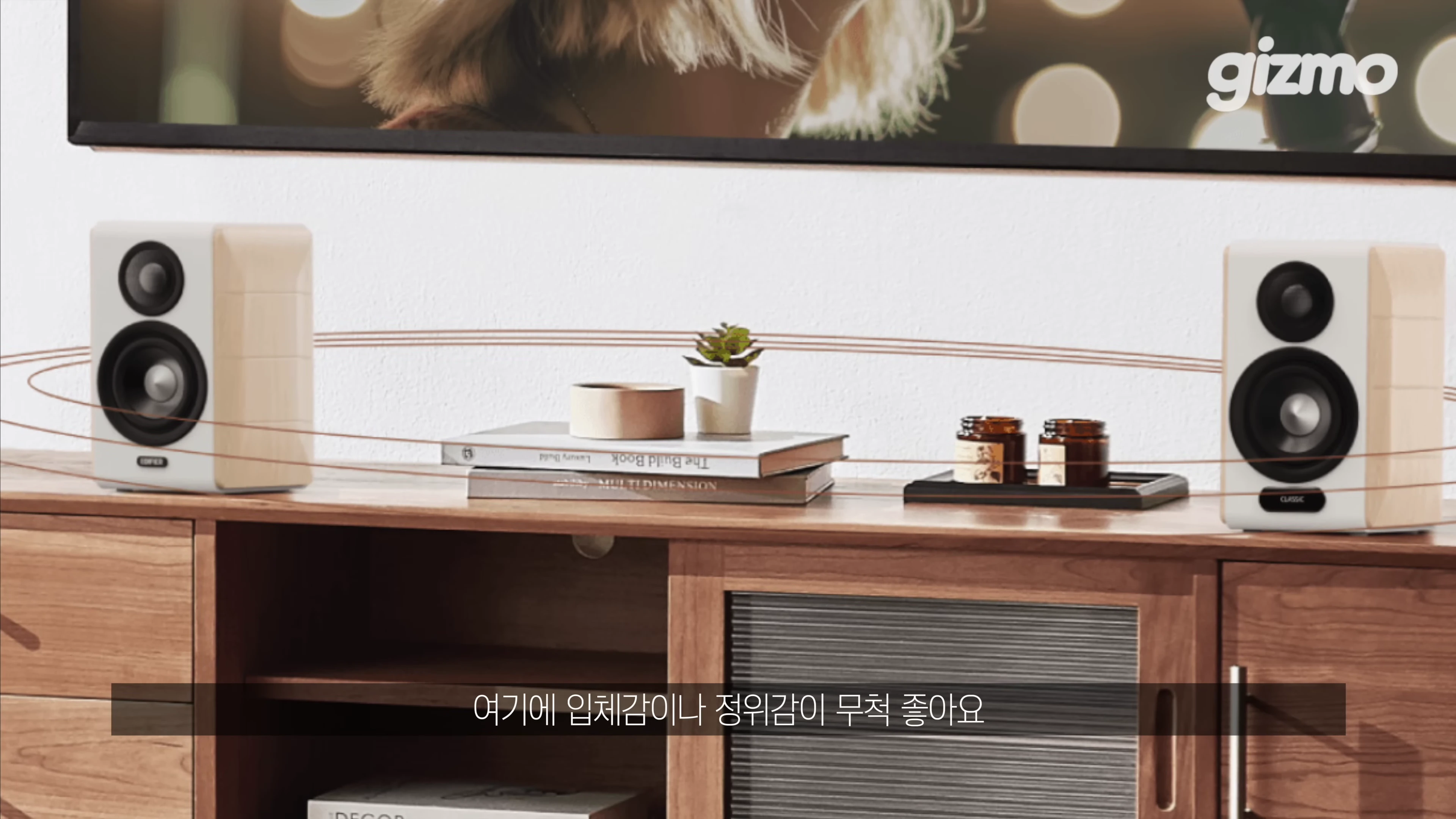 나무 무늬와 흰색이 조화로운 2채널 스피커 한 쌍이 거실장 위에 놓여 있고, 그 주위로 소리의 입체감을 나타내는 원형 그래픽이 그려져 있습니다.