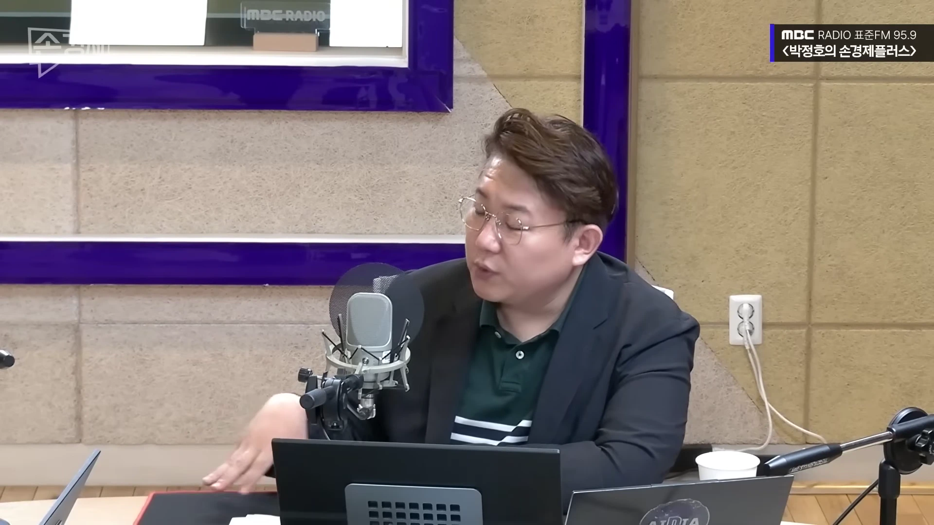 안경을 쓴 남성이 라디오 스튜디오에서 마이크 앞에 앉아 이야기를 나누고 있다.