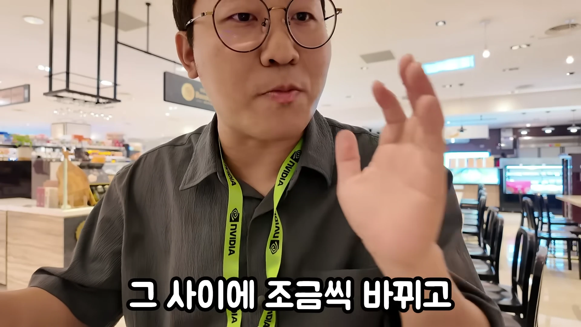 엔비디아 로고가 새겨진 목걸이를 착용한 안경 쓴 남성이 실내에서 손짓하며 이야기하고 있다.