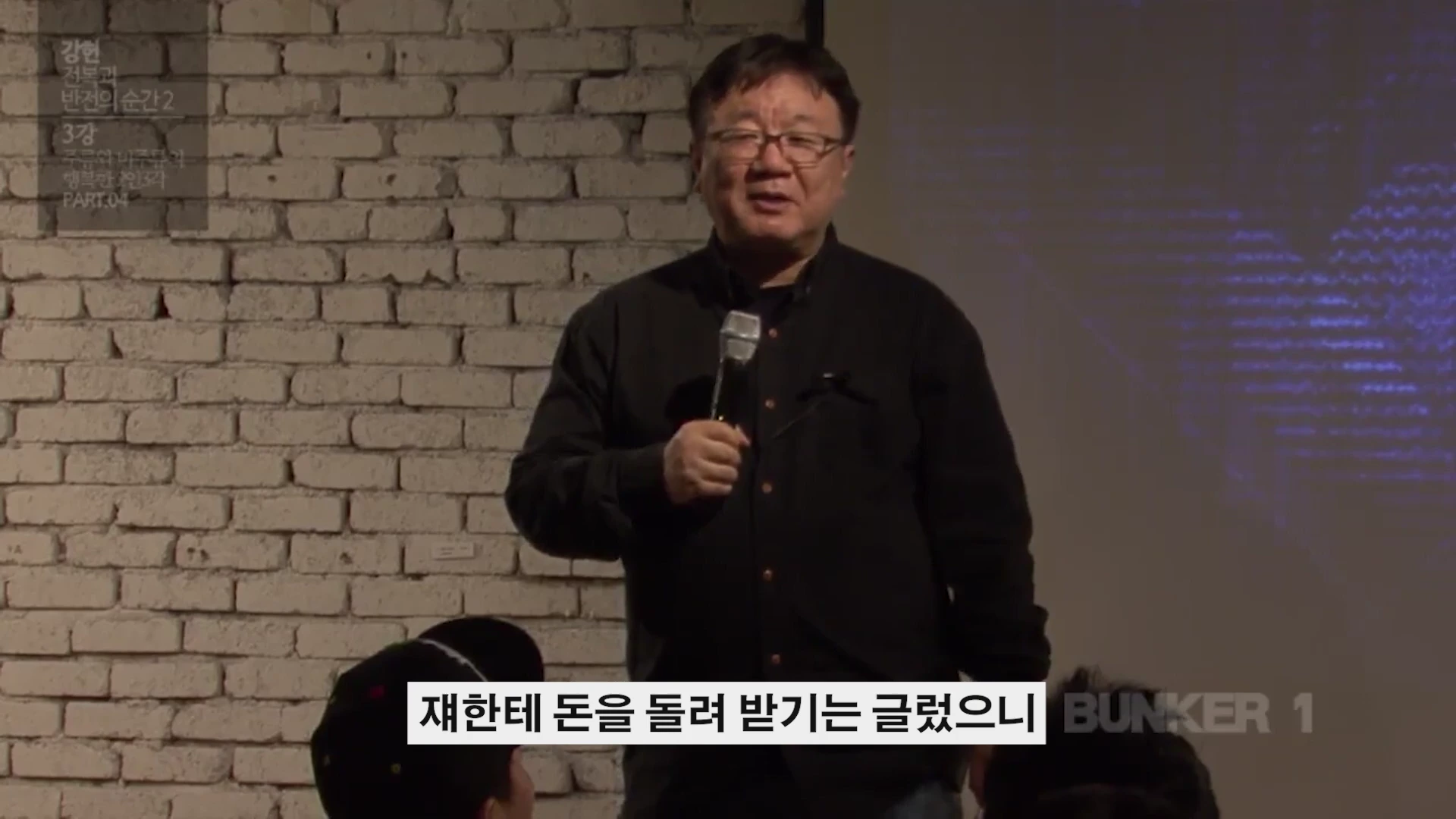 흰 벽돌 배경 앞에서 마이크를 들고 강연하는 중년 남성