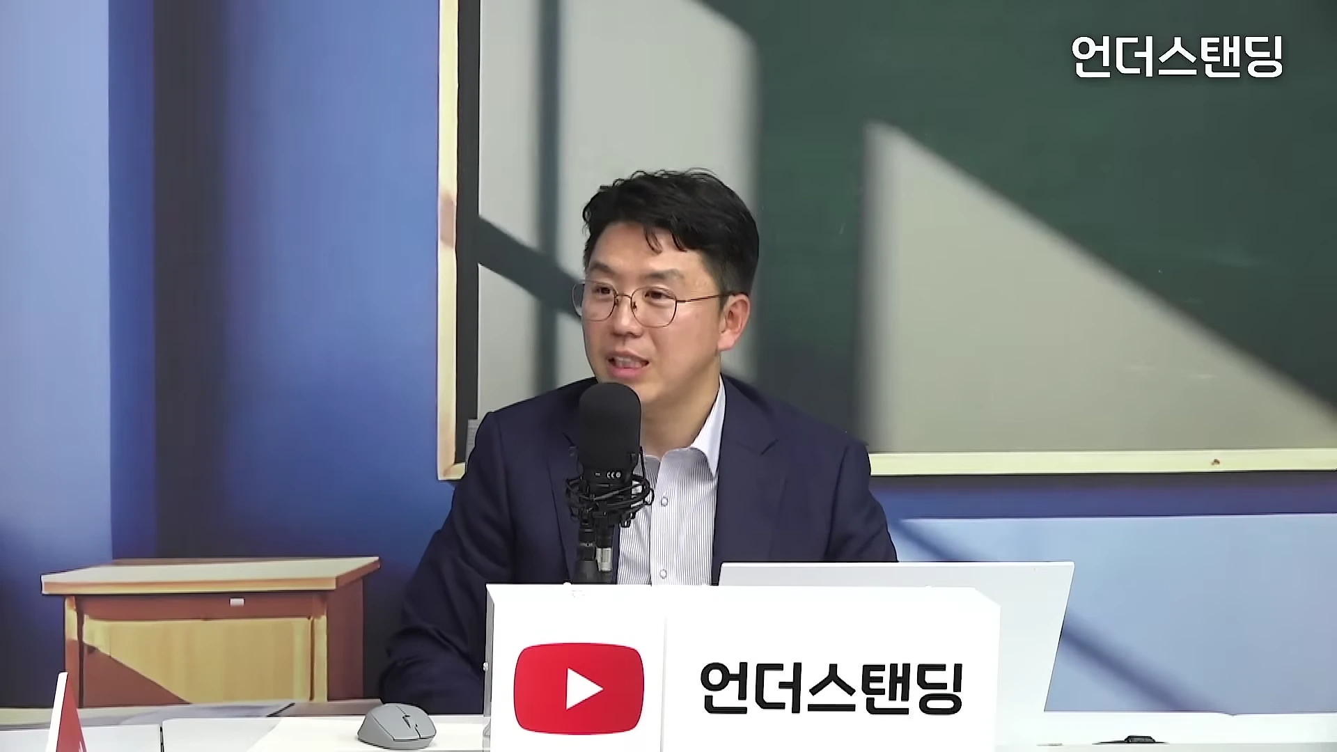 안경을 쓴 남성이 스튜디오에서 마이크 앞에 앉아 이야기를 나누고 있다.