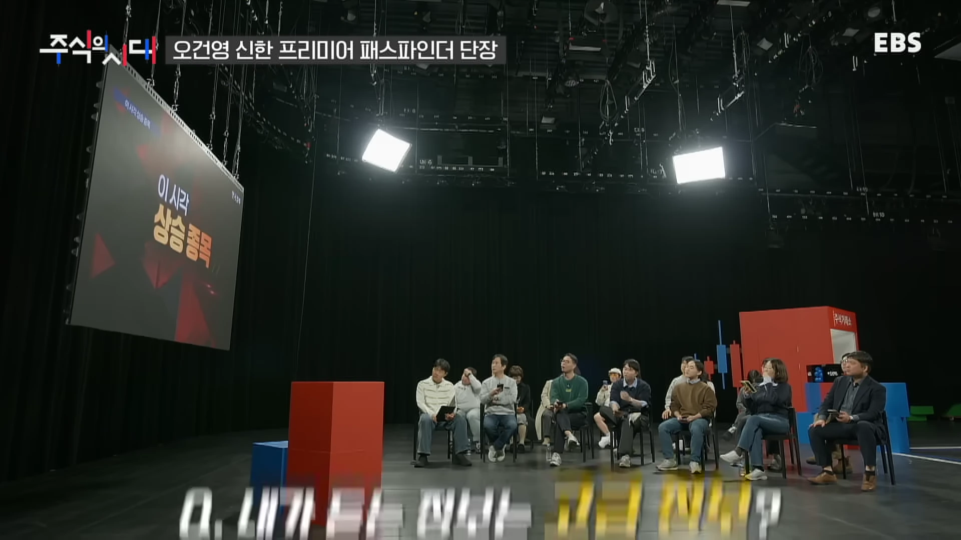 어두운 스튜디오에서 사람들이 의자에 앉아 대형 화면을 바라보고 있는 모습