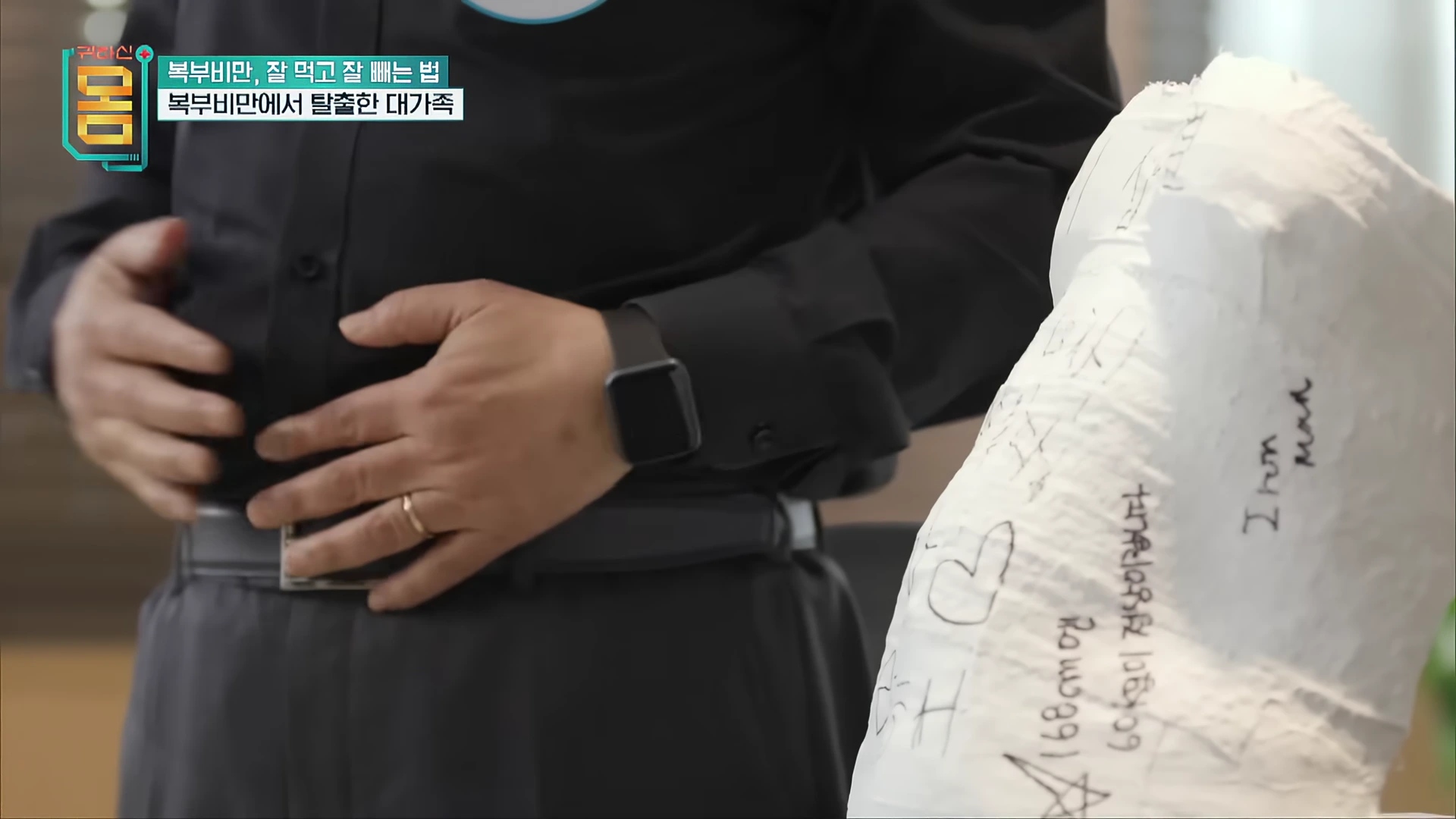 검은 셔츠를 입은 남성이 자신의 허리 부근에 손을 올리고 있고, 옆에는 글씨가 적힌 흰색 석고 모형이 놓여 있다.