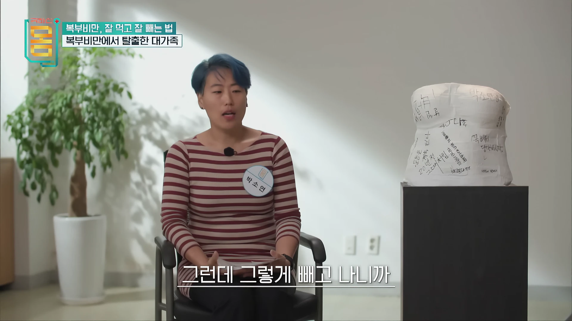 줄무늬 상의를 입은 여성이 의자에 앉아 인터뷰하고 있으며, 옆에는 글귀가 적힌 복부 모형이 놓여 있다.
