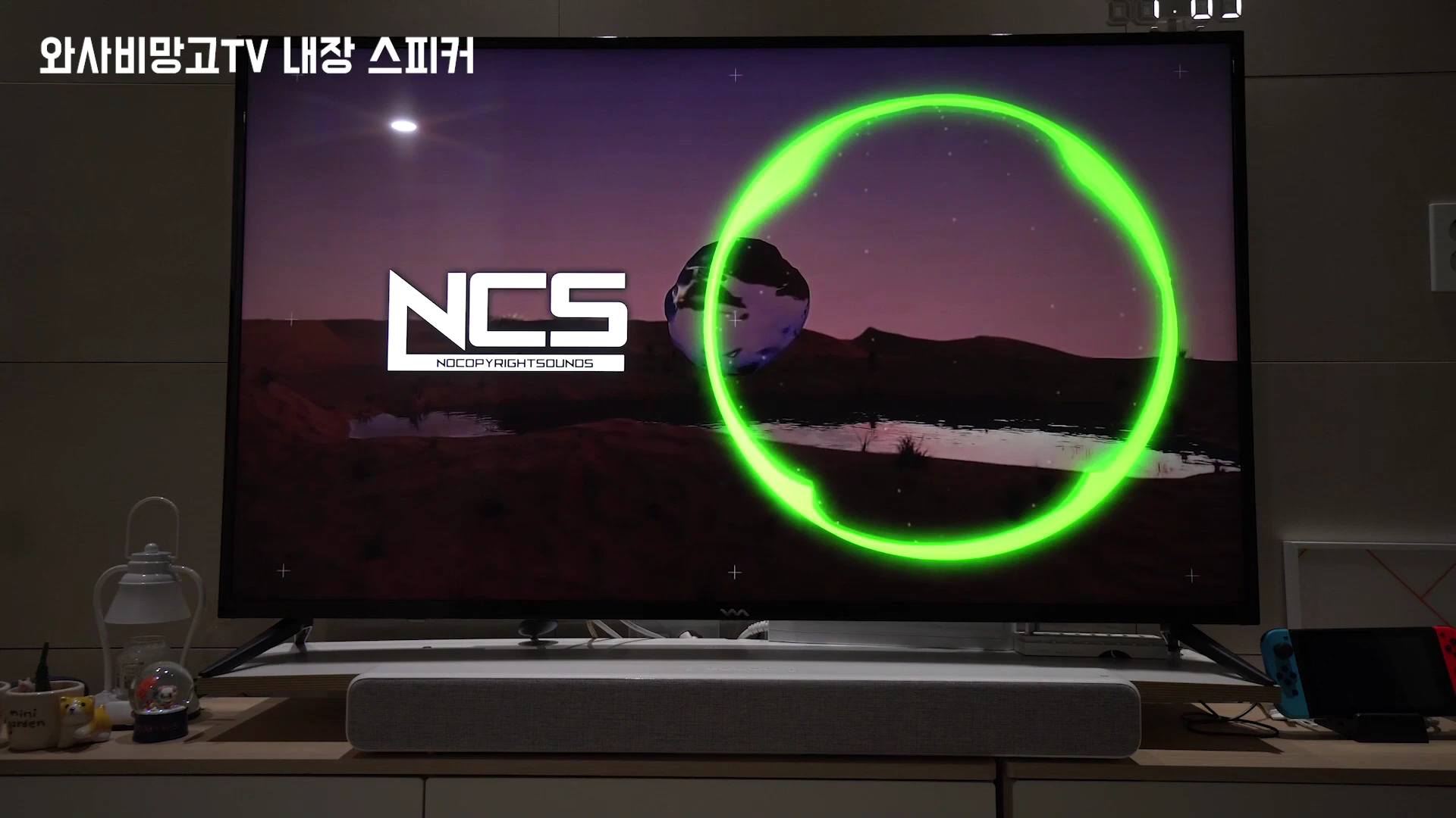 거실에 놓인 55인치 TV 화면에 NCS 로고와 녹색 원형 파형이 표시되어 있고, 아래에는 사운드바가 배치된 모습