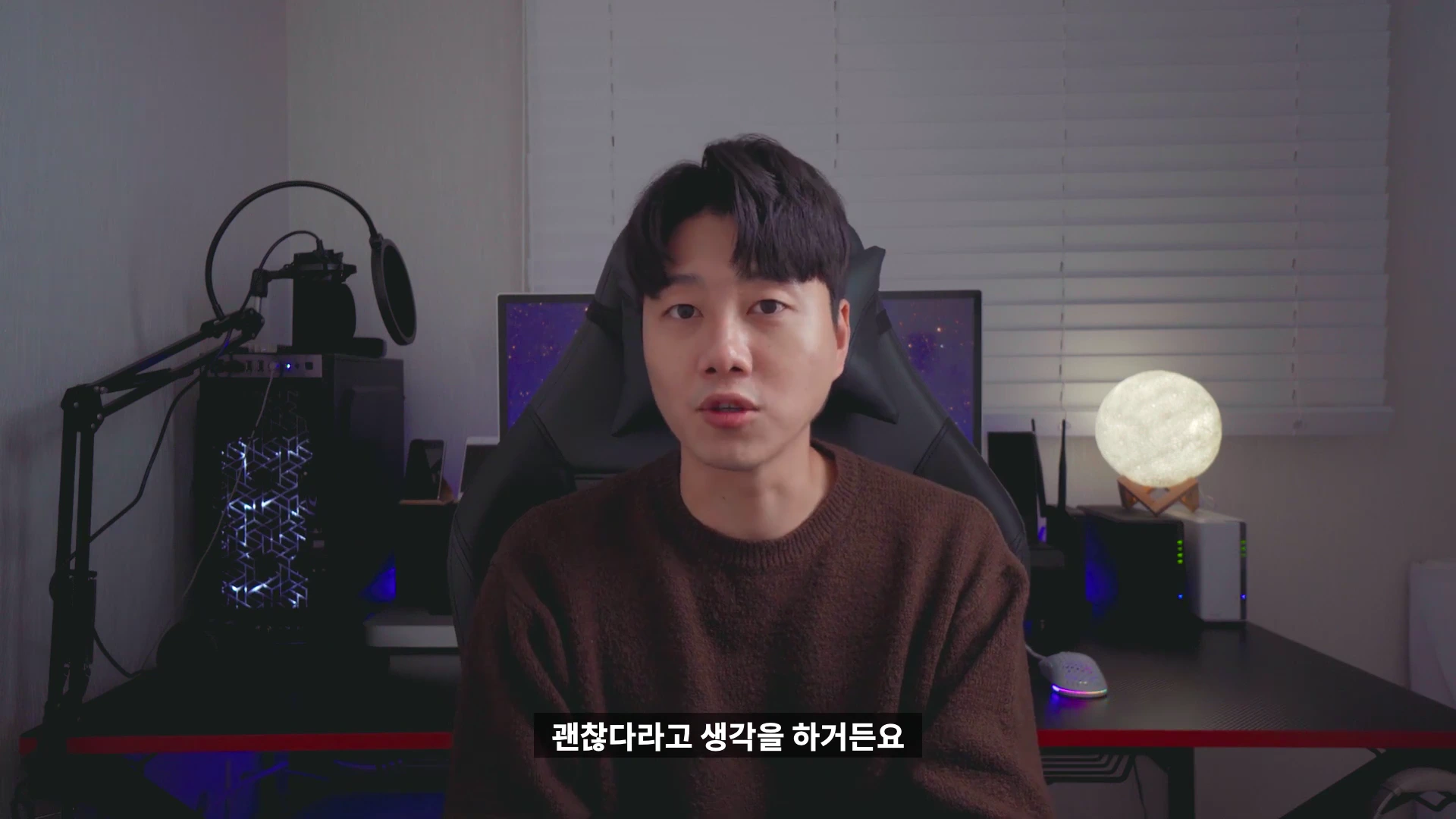 어두운 방 안에서 갈색 니트를 입은 남성이 의자에 앉아 정면을 바라보며 이야기하고 있다.