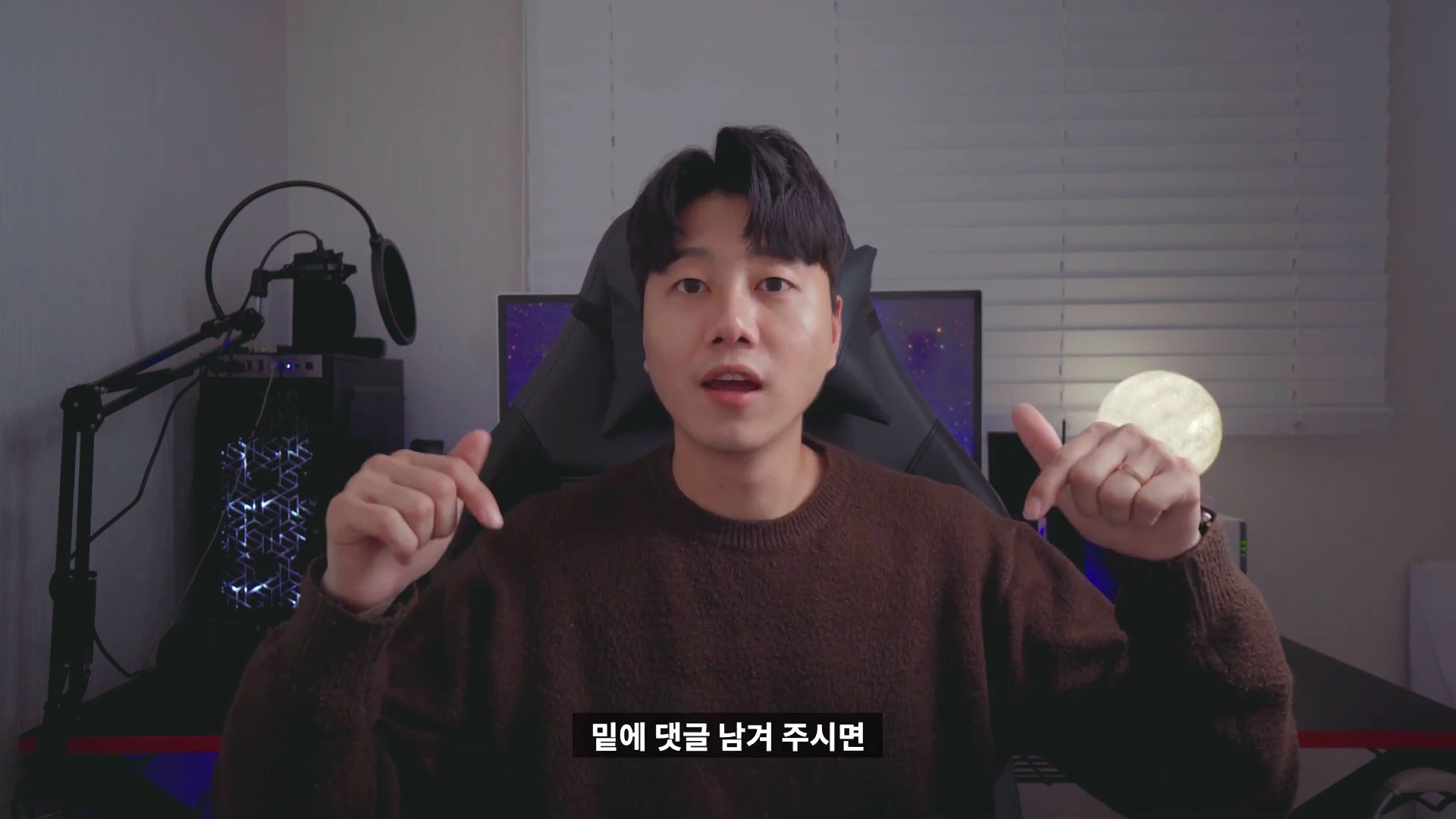 갈색 니트를 입은 남성이 책상 앞에 앉아 양손으로 아래를 가리키며 이야기하고 있다.