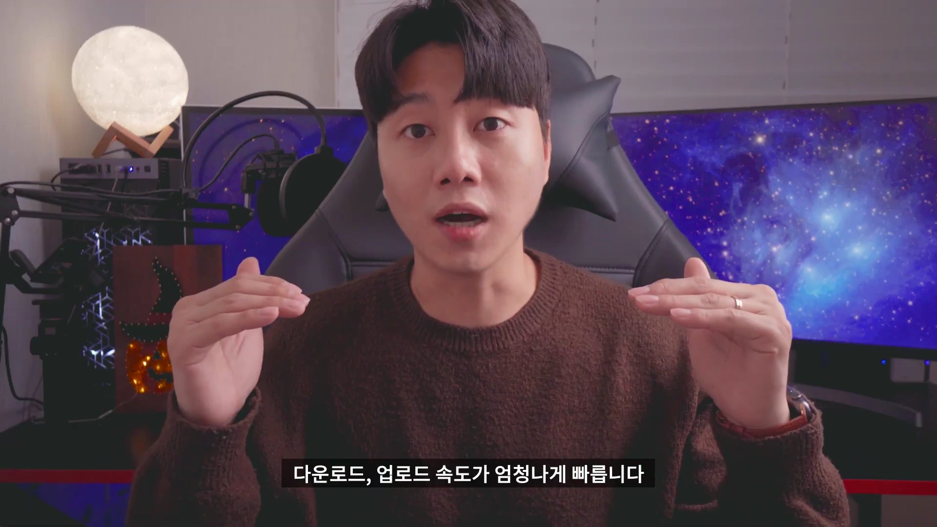 갈색 니트를 입은 남성이 스튜디오 의자에 앉아 양손으로 높이를 비교하며 설명하고 있는 모습