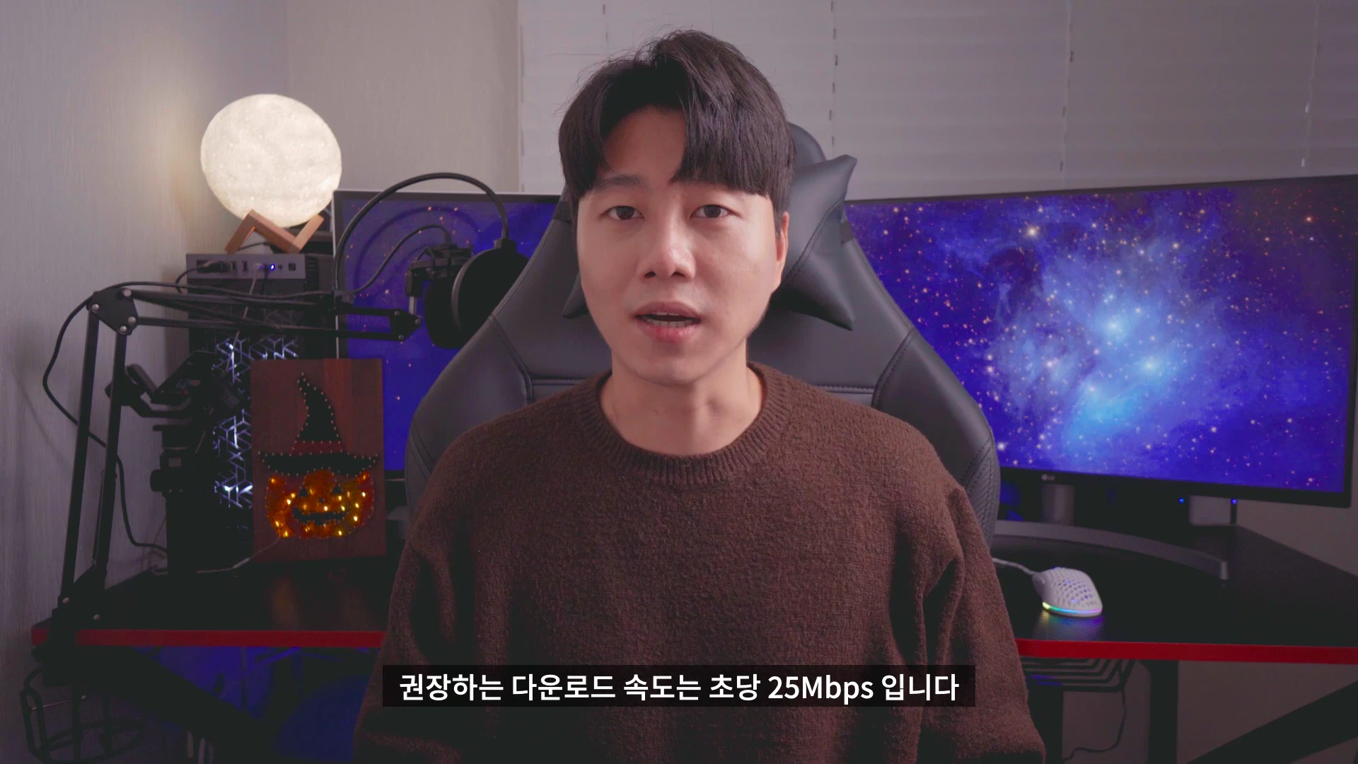 갈색 니트를 입은 남성이 게이밍 의자에 앉아 카메라를 응시하며 이야기하고 있다.