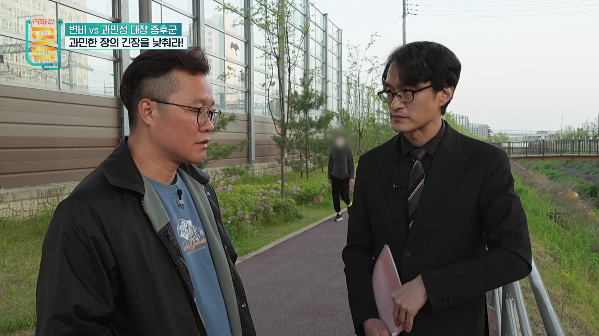야외 산책로에서 안경을 쓴 두 남성이 진지하게 대화를 나누고 있는 모습