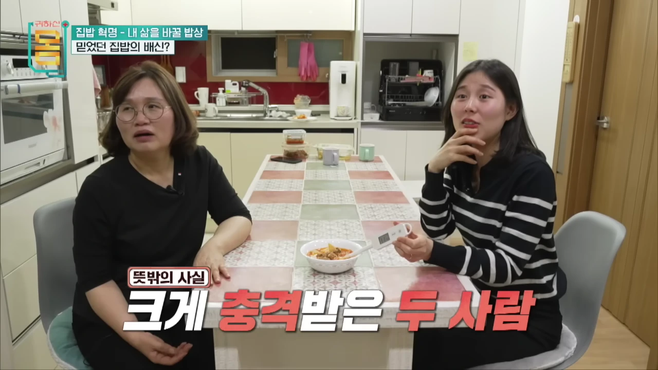 주방 식탁에 마주 앉은 두 여성이 염도계로 김치찌개 국물을 측정하며 놀란 표정을 짓고 있다.