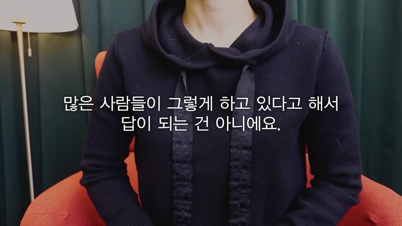 검은색 후드티를 입은 사람이 의자에 앉아 있고 화면 중앙에 자막이 떠 있다.