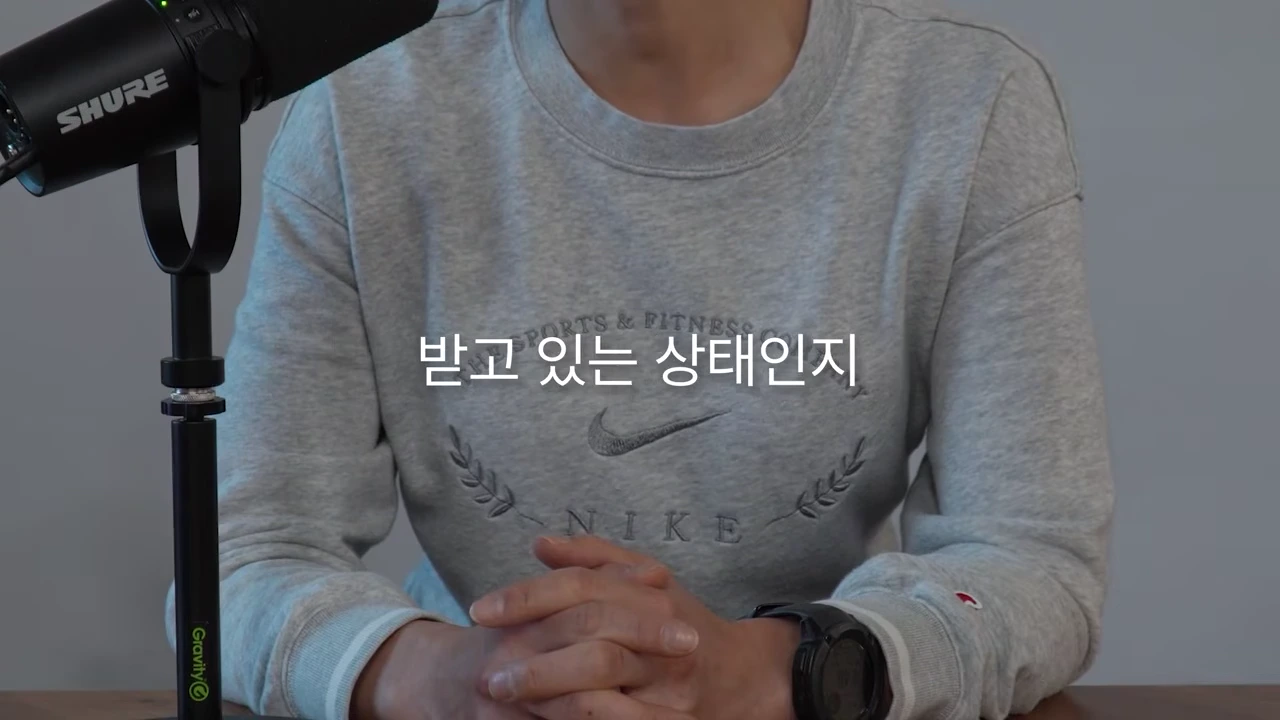 회색 맨투맨을 입은 사람이 마이크 앞에 앉아 두 손을 모으고 있는 모습