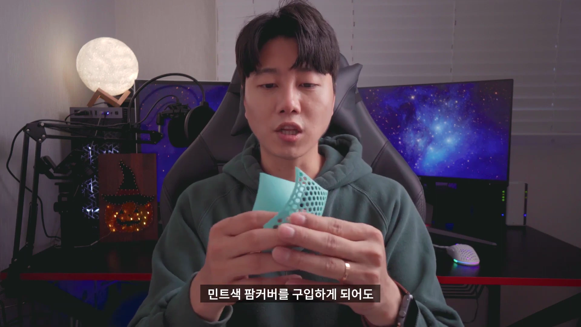 게이밍 룸을 배경으로 한 남성이 민트색 타공 마우스 커버를 손에 들고 설명하는 모습