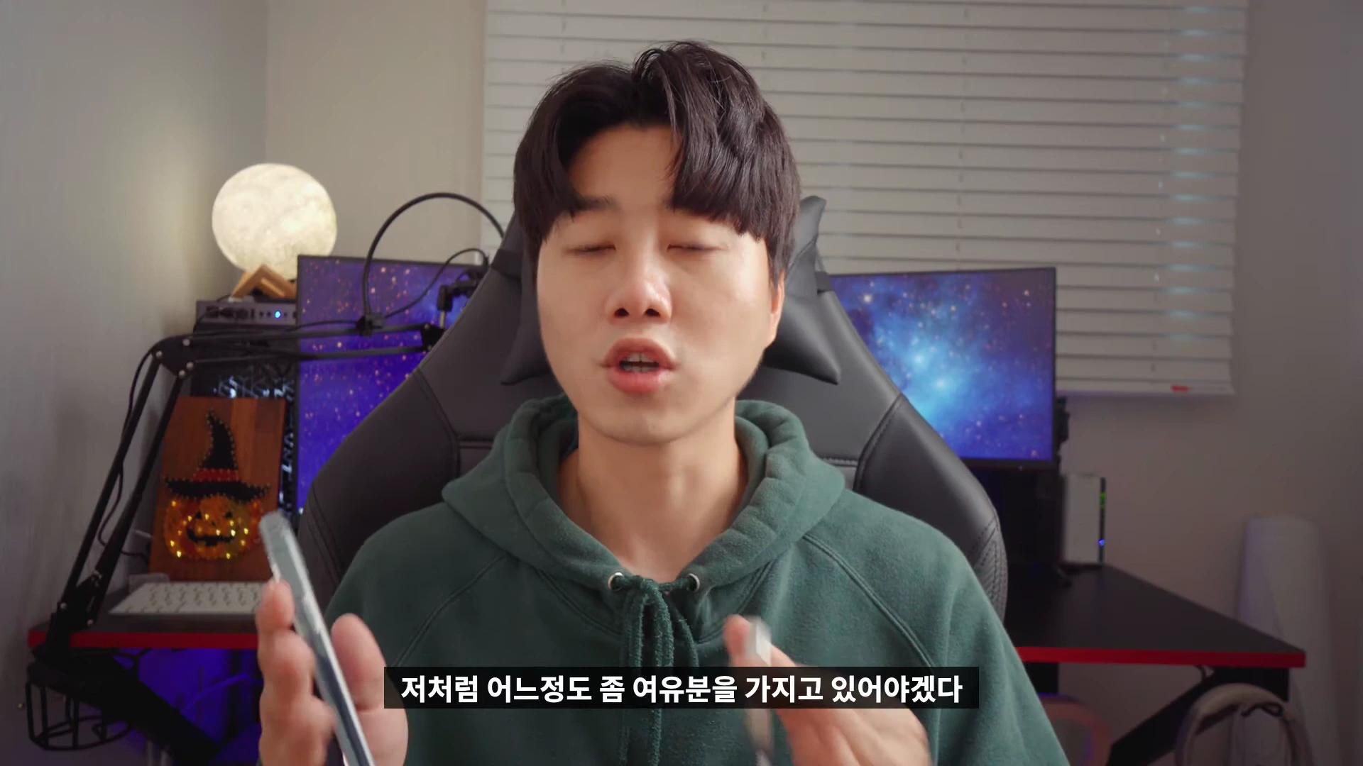 초록색 후드티를 입은 남성이 방 안에서 스마트폰 케이스를 들고 설명하고 있다.
