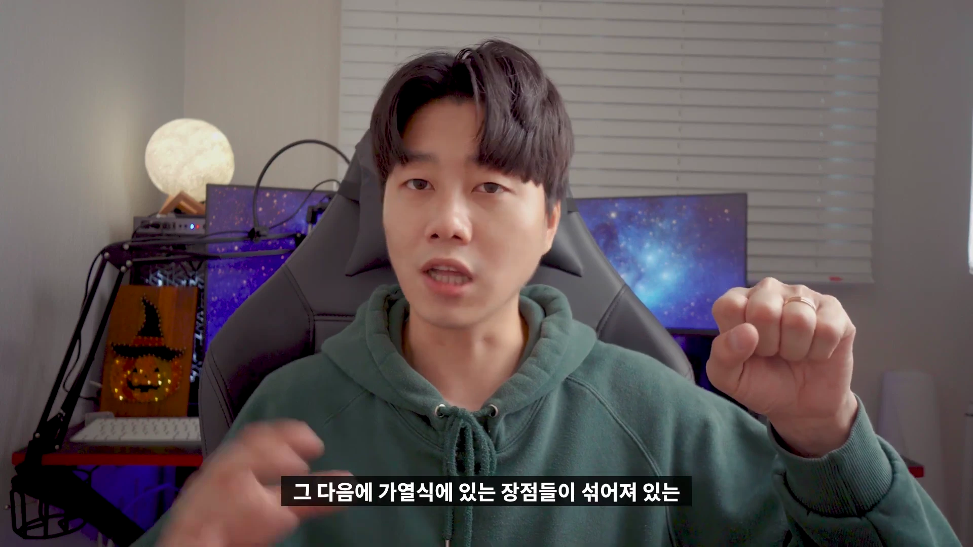 초록색 후드티를 입은 남성이 방 안에서 손짓하며 이야기하고 있는 모습