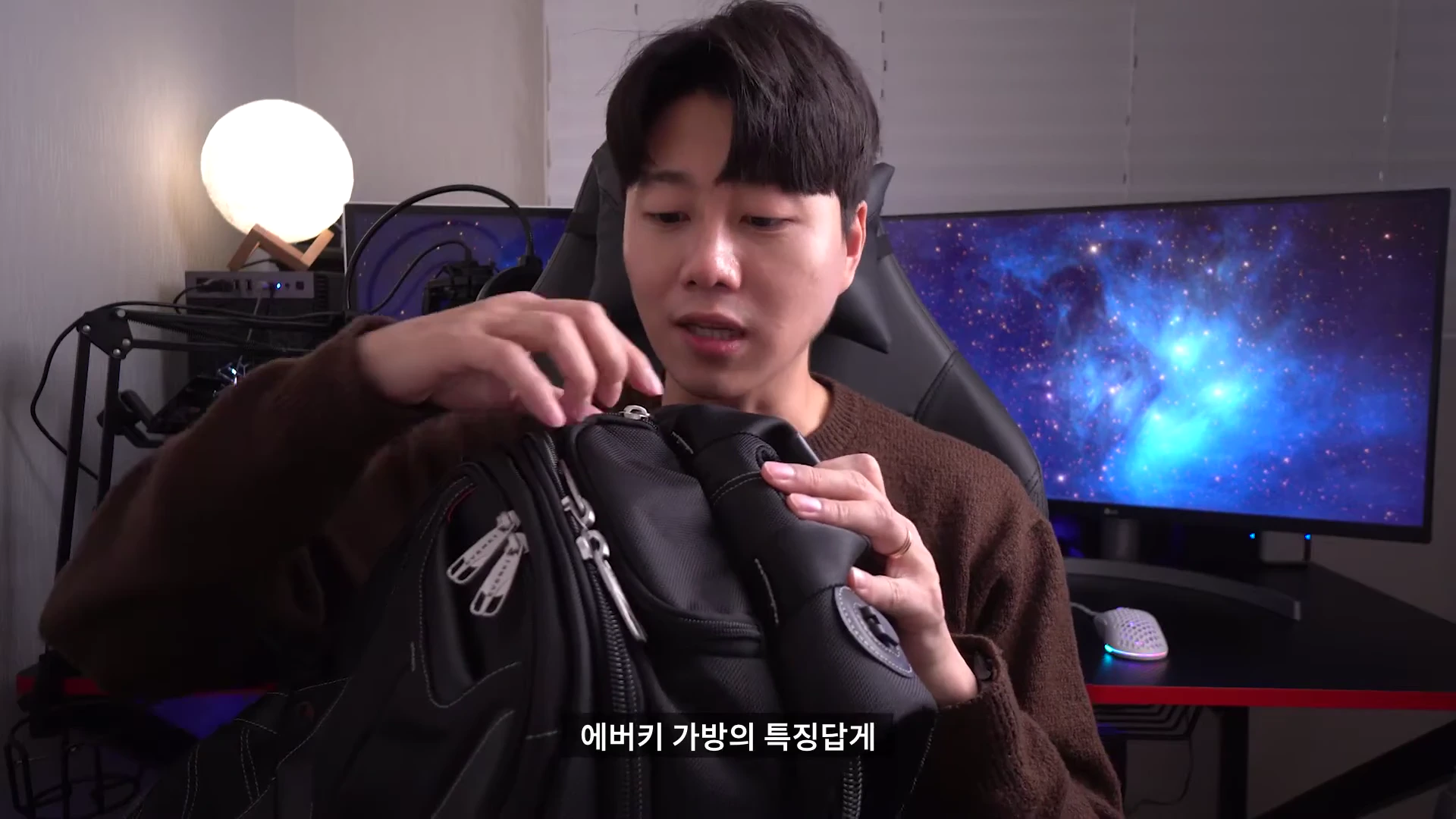 갈색 니트를 입은 남성이 책상에 앉아 검은색 백팩의 상단 포켓을 열어보고 있는 모습