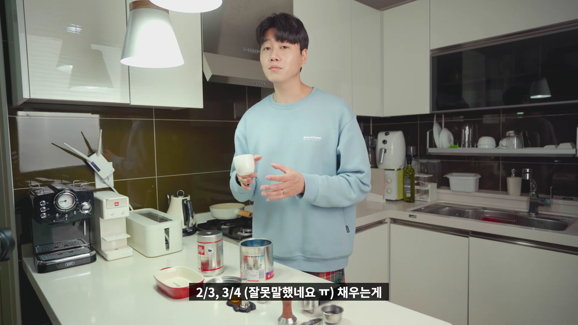 주방에서 하늘색 맨투맨을 입은 남성이 작은 에스프레소 잔을 들고 설명하는 모습