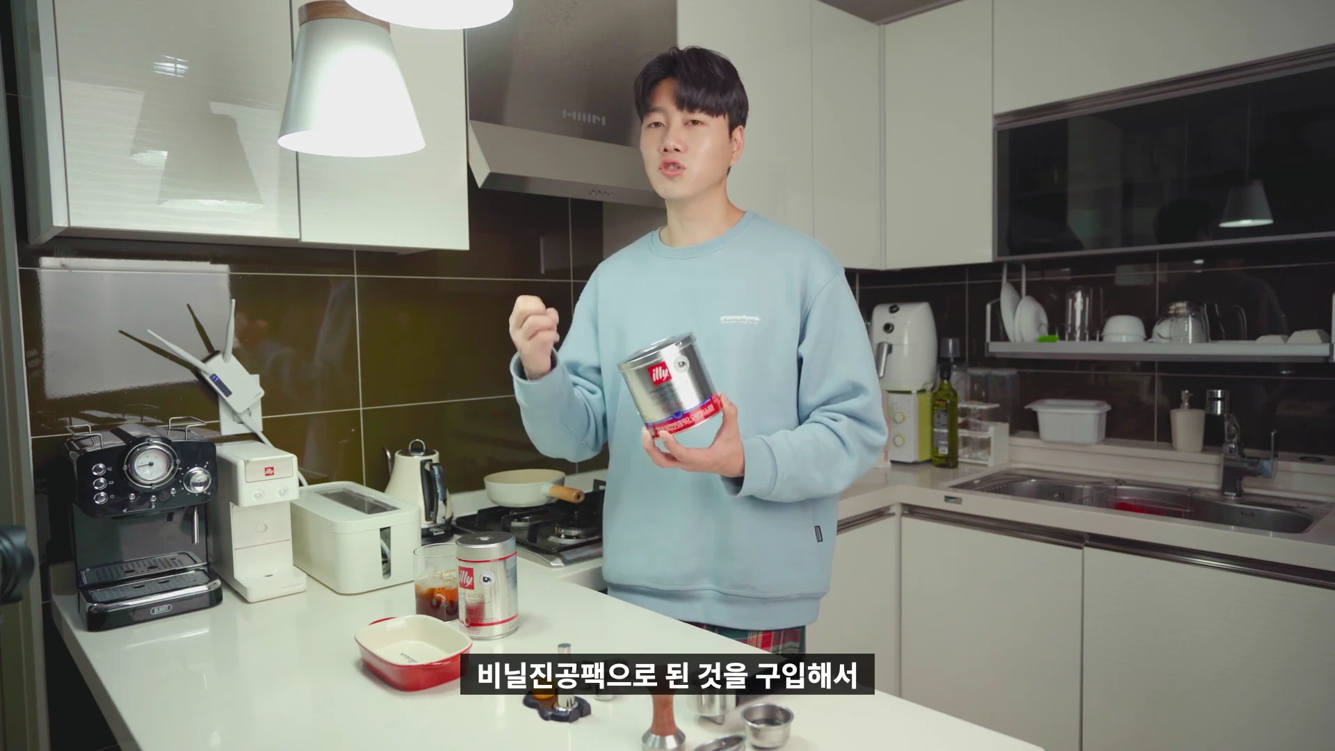 주방에서 하늘색 맨투맨을 입은 남성이 일리 커피 캔을 들고 설명하는 모습