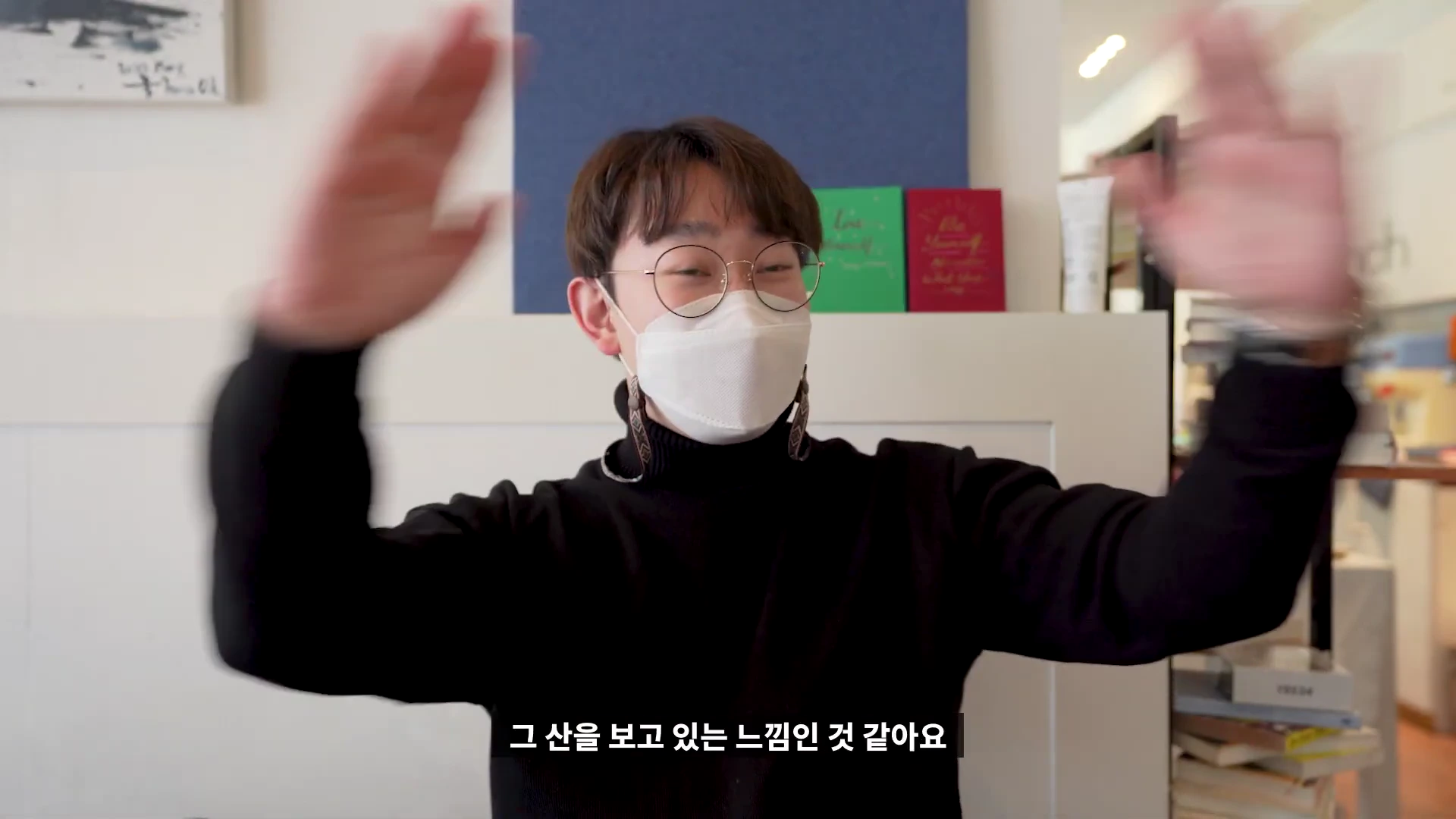 안경을 쓰고 마스크를 착용한 남성이 실내에서 양손을 들어 올리며 인사하는 모습