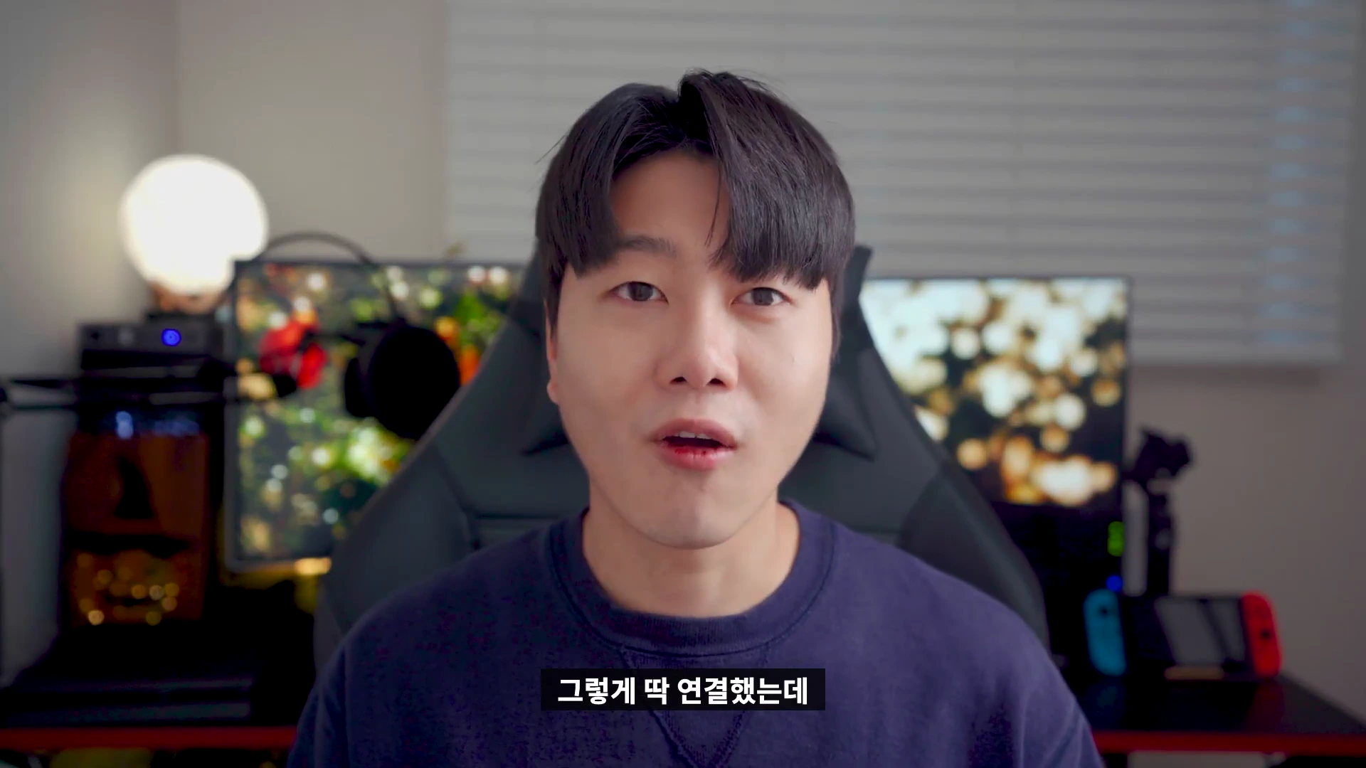 어두운 방 안에서 게이밍 의자에 앉아 카메라를 정면으로 응시하며 이야기하는 남성