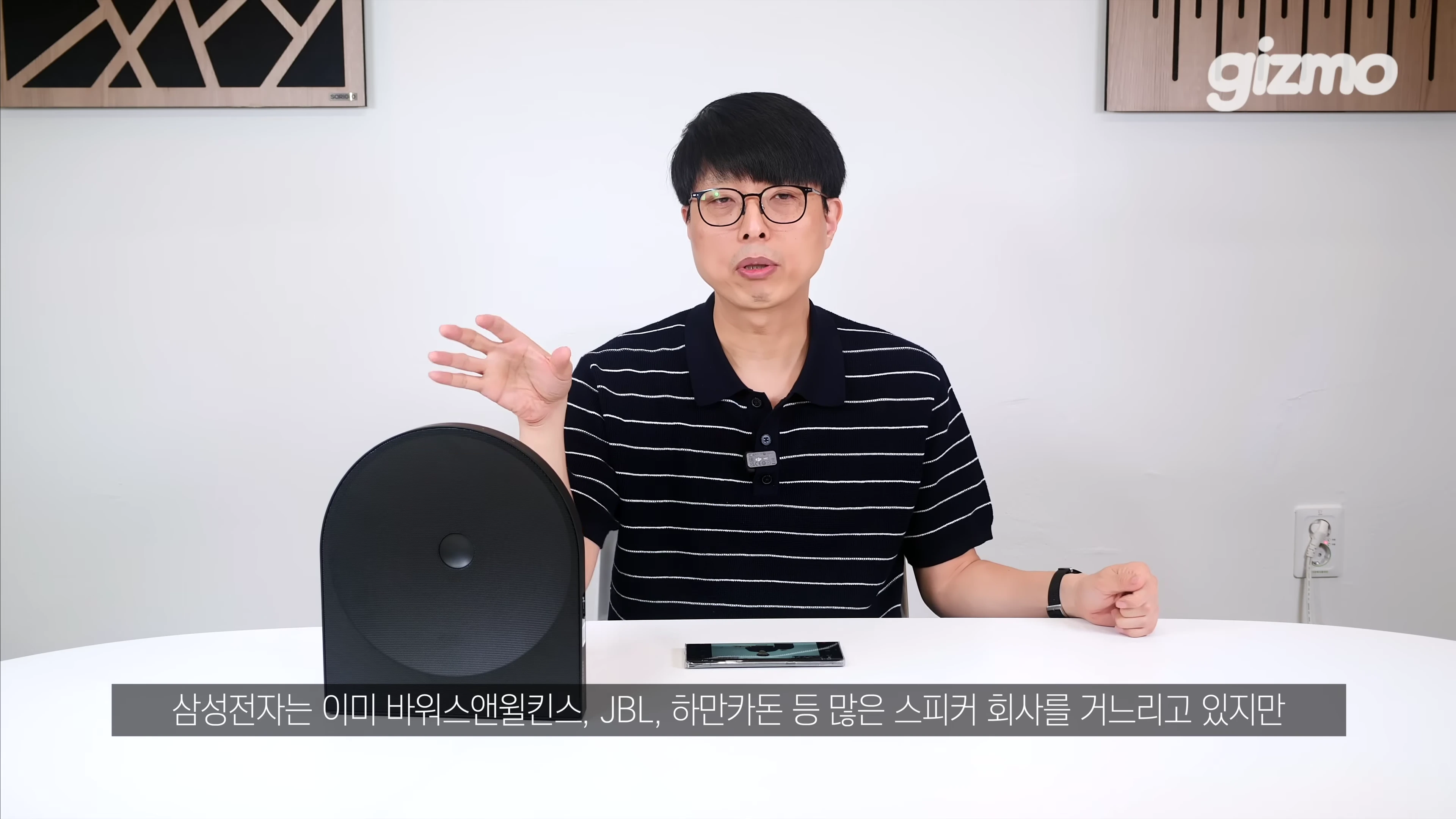 안경을 쓴 남성이 흰색 테이블 위에 놓인 검은색 원형 스피커 옆에서 손짓하며 설명하고 있다.