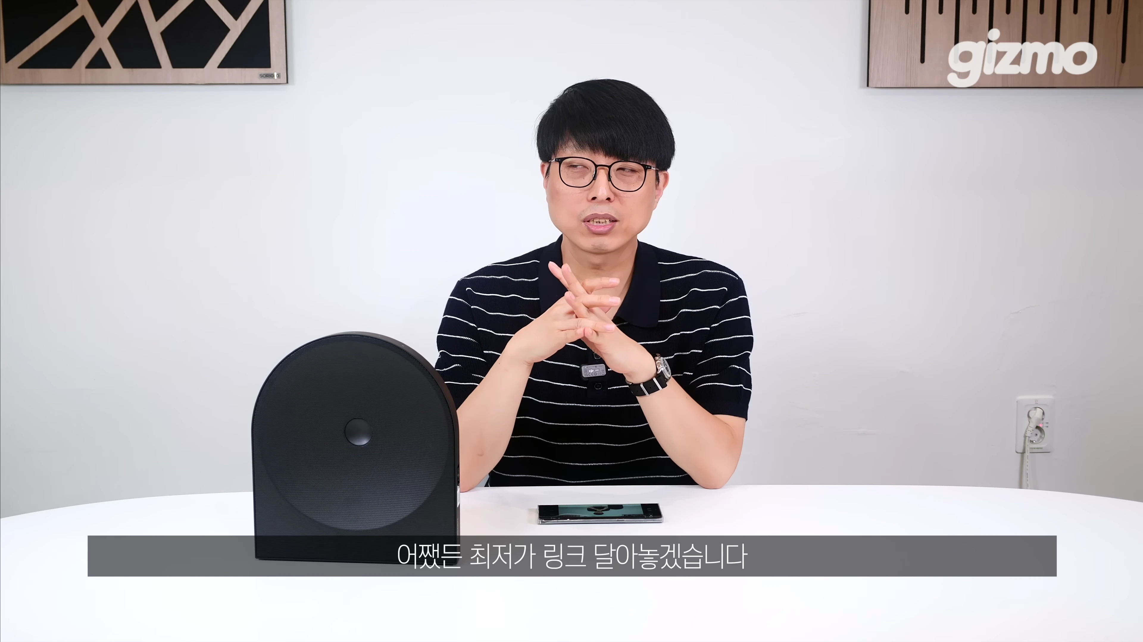 안경을 쓴 남성이 책상 앞에 앉아 검은색 원형 스피커를 앞에 두고 이야기하는 모습
