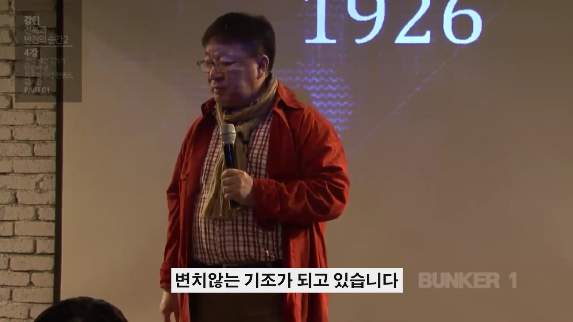 강연장에서 마이크를 들고 서 있는 남성 뒤로 1926이라는 숫자가 적힌 스크린이 보인다.