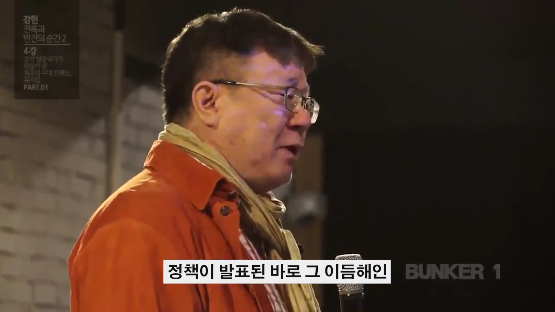 주황색 재킷을 입고 안경을 쓴 남성이 마이크를 잡고 강연하는 모습