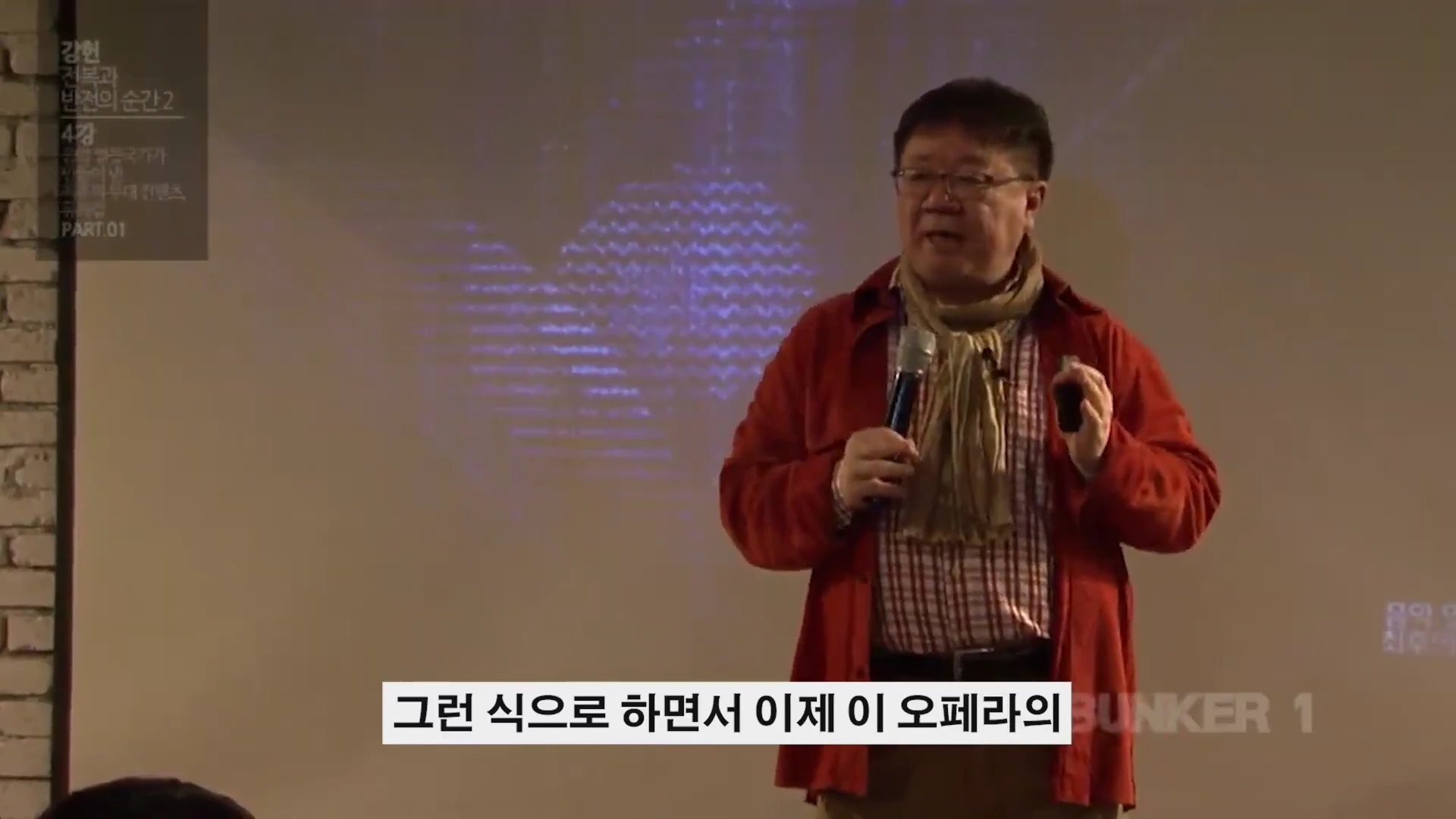 붉은 셔츠를 입은 중년 남성이 마이크를 들고 강연하는 모습