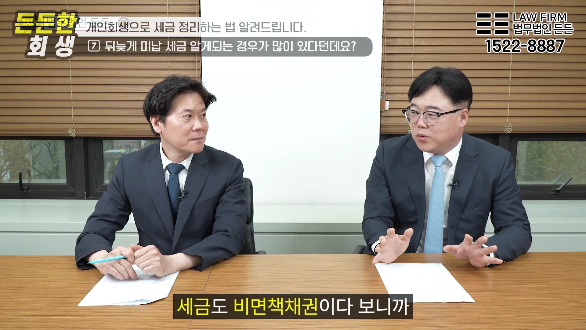 정장을 입은 두 남성이 책상에 앉아 대화를 나누고 있는 스튜디오 인터뷰 장면