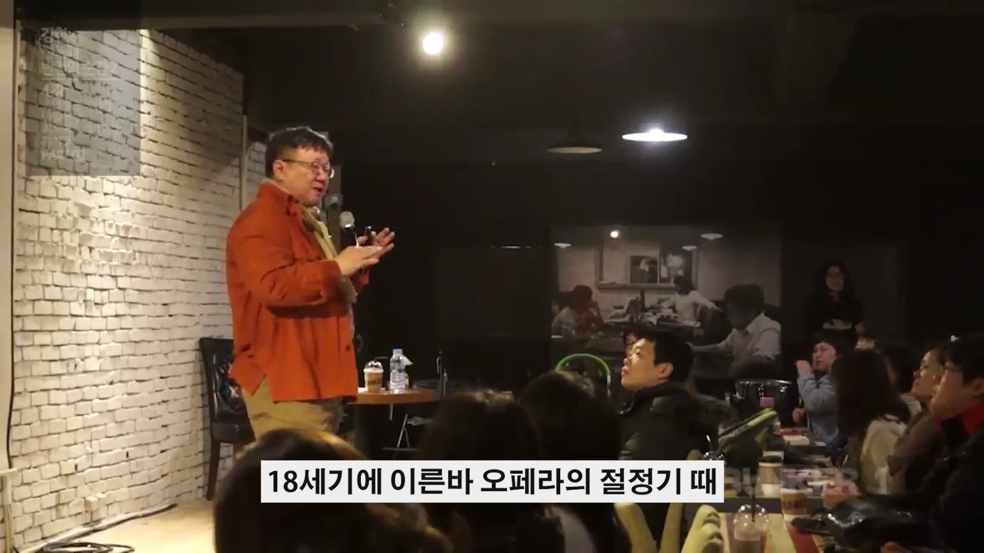 흰 벽돌 배경의 무대 위에서 주황색 셔츠를 입은 남성이 마이크를 들고 청중을 향해 강연하고 있다.