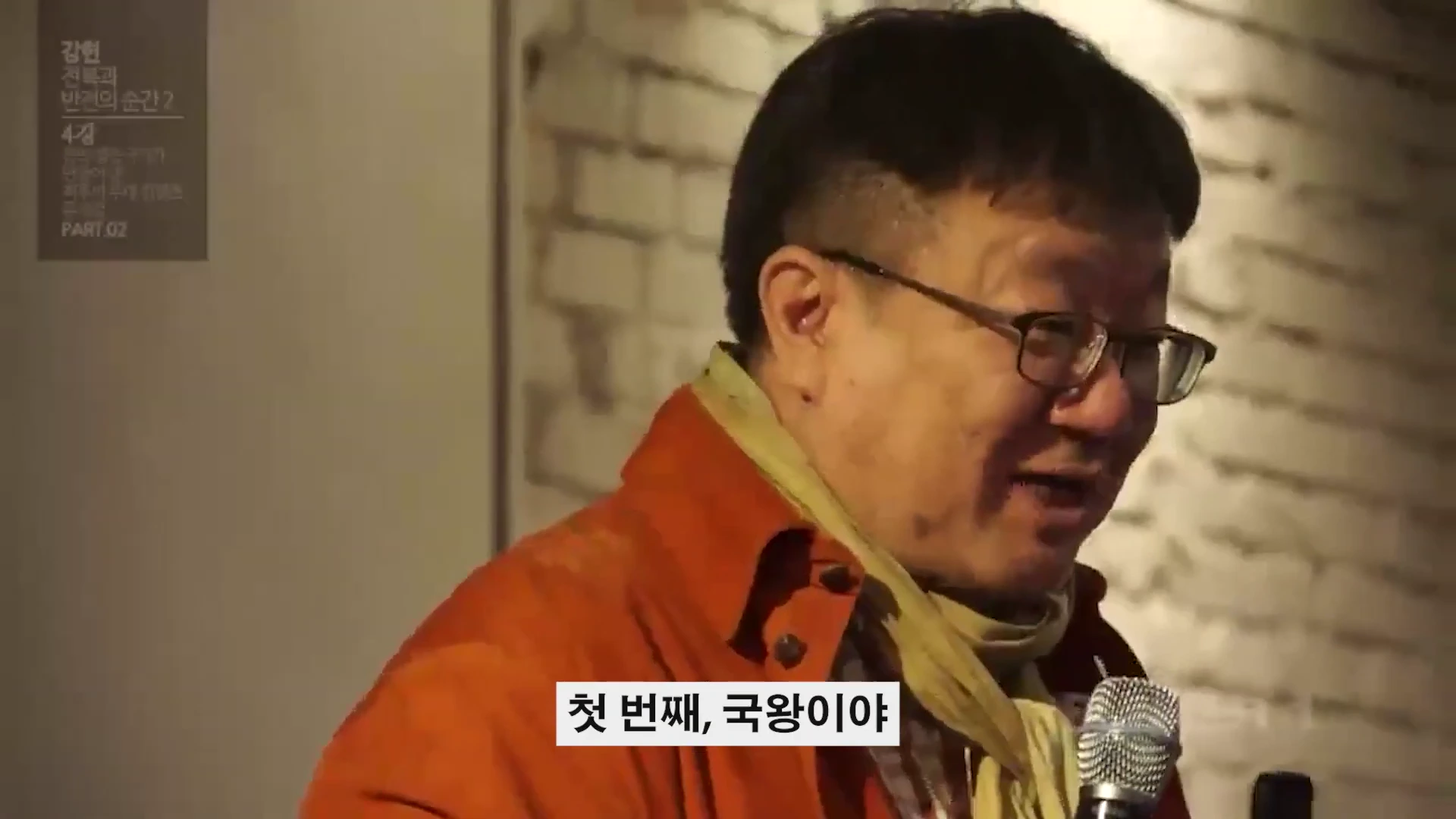 안경을 쓴 중년 남성이 주황색 재킷을 입고 마이크를 든 채 강연하는 모습