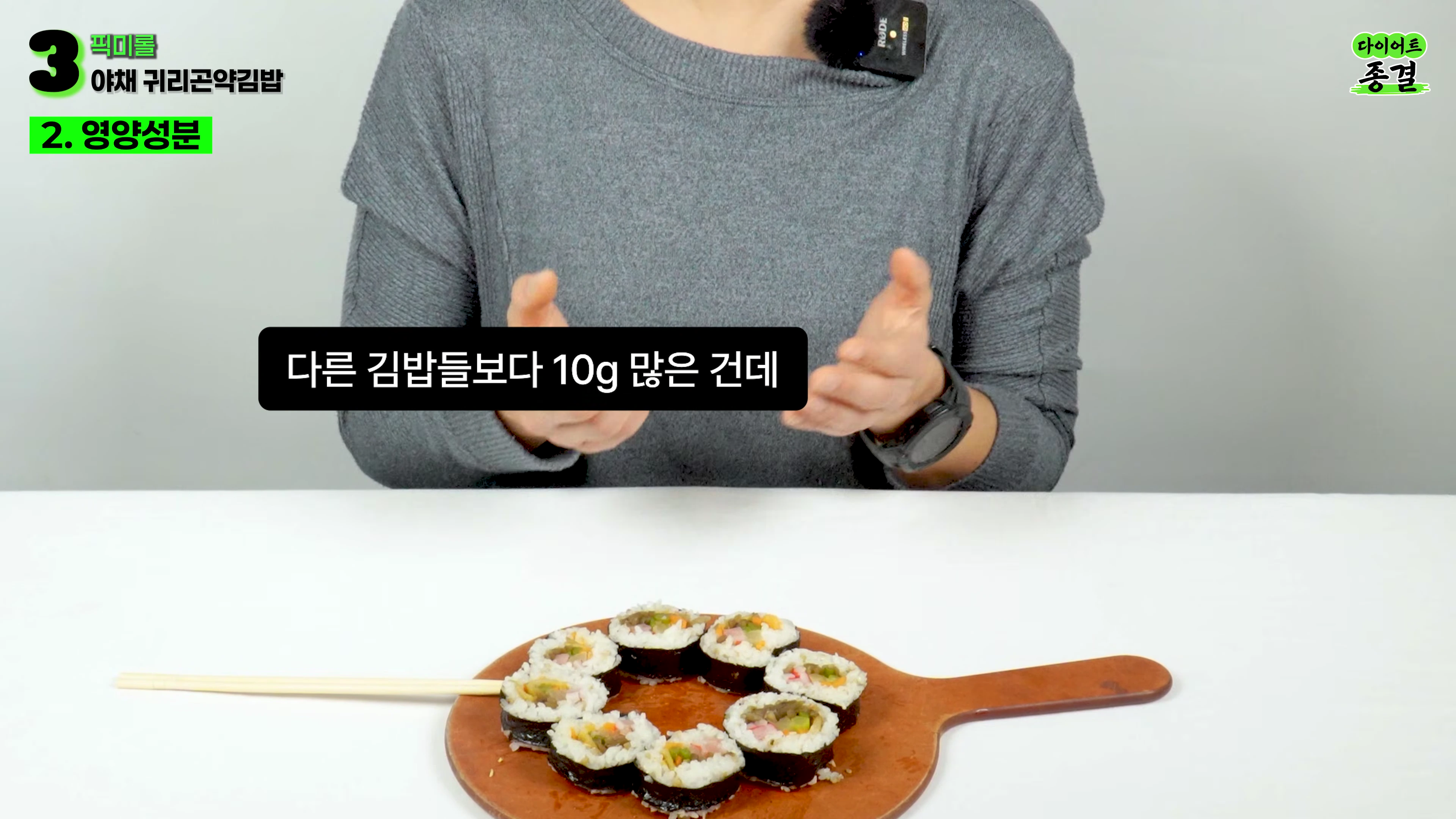 회색 니트를 입은 사람이 테이블 위에 놓인 김밥 접시를 앞에 두고 손짓하며 설명하는 모습