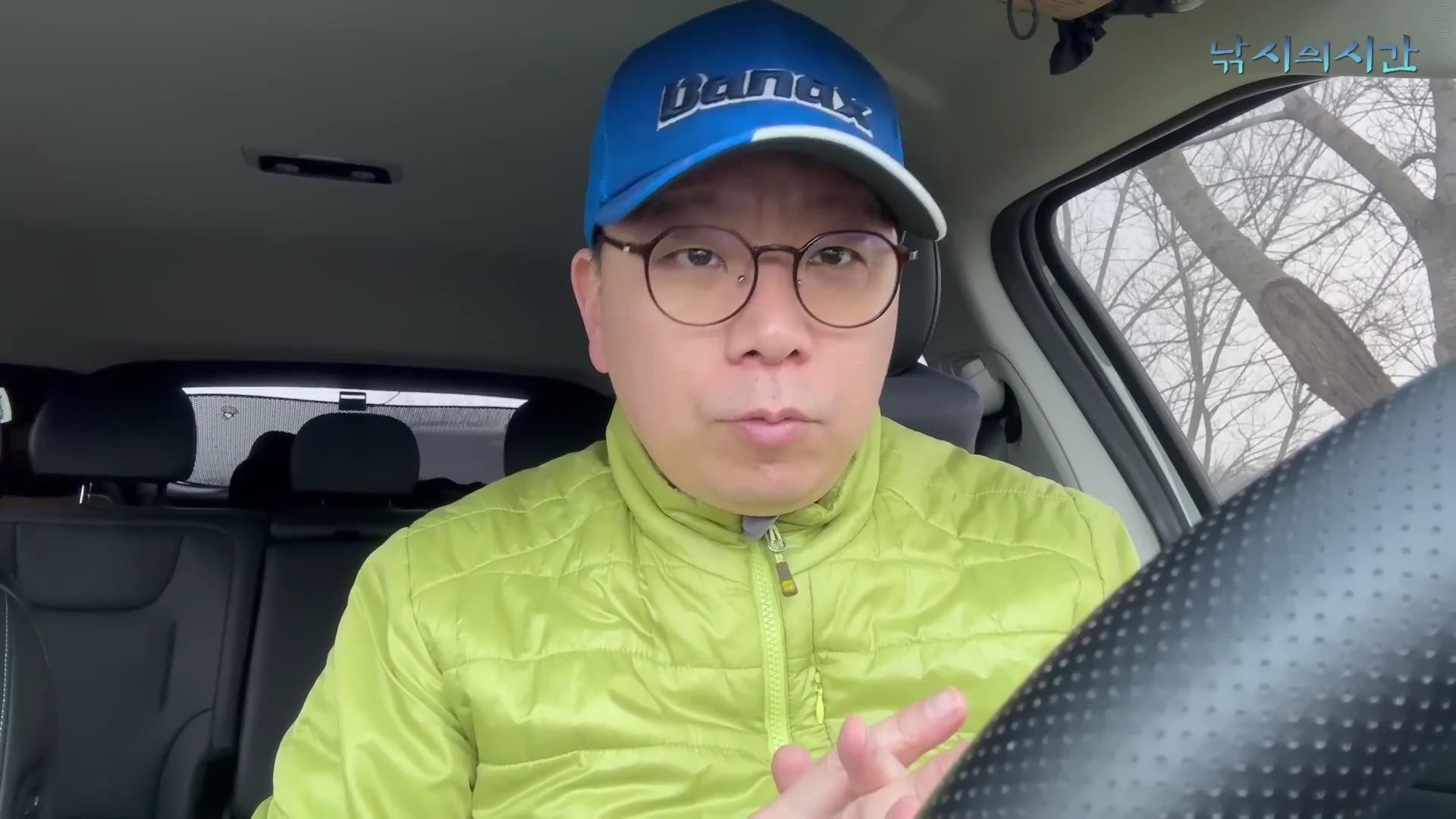 파란색 모자와 안경을 쓴 남성이 자동차 운전석에 앉아 정면을 바라보며 이야기하고 있다.