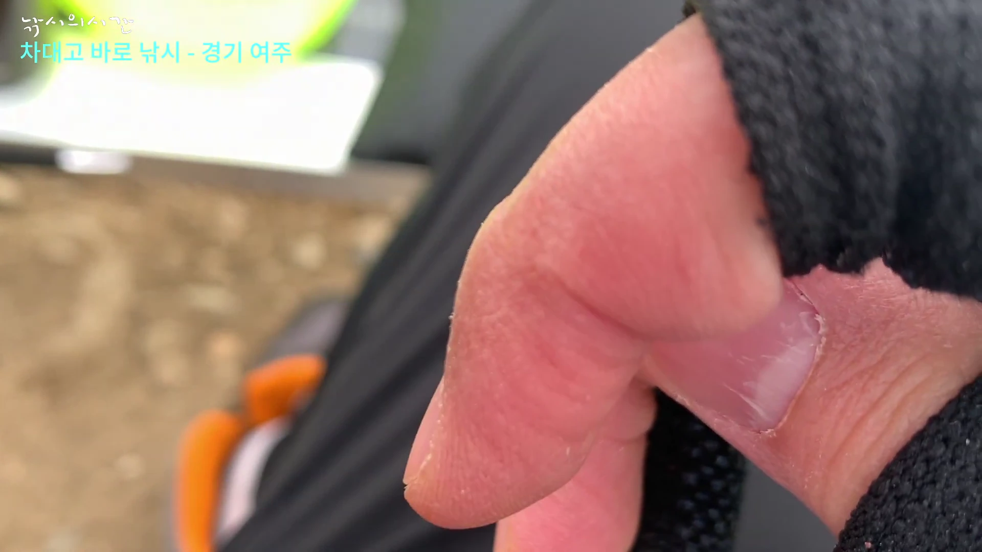 낚시 장갑을 낀 손이 화면 가까이 보이고 배경으로 흐릿한 야외 낚시터가 나타나는 영상 캡처 화면