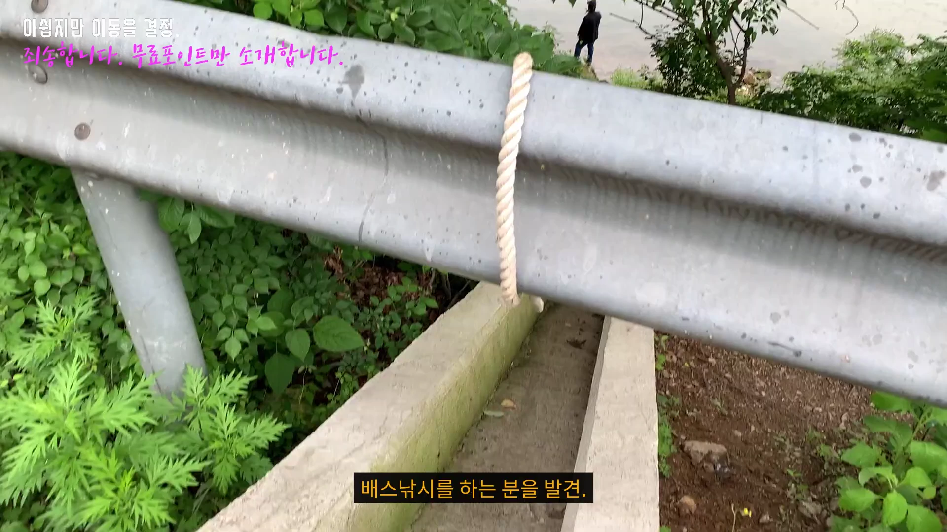 도로 가드레일에 묶인 밧줄과 그 아래로 내려가는 좁은 비탈길, 멀리 낚시하는 사람이 보인다.