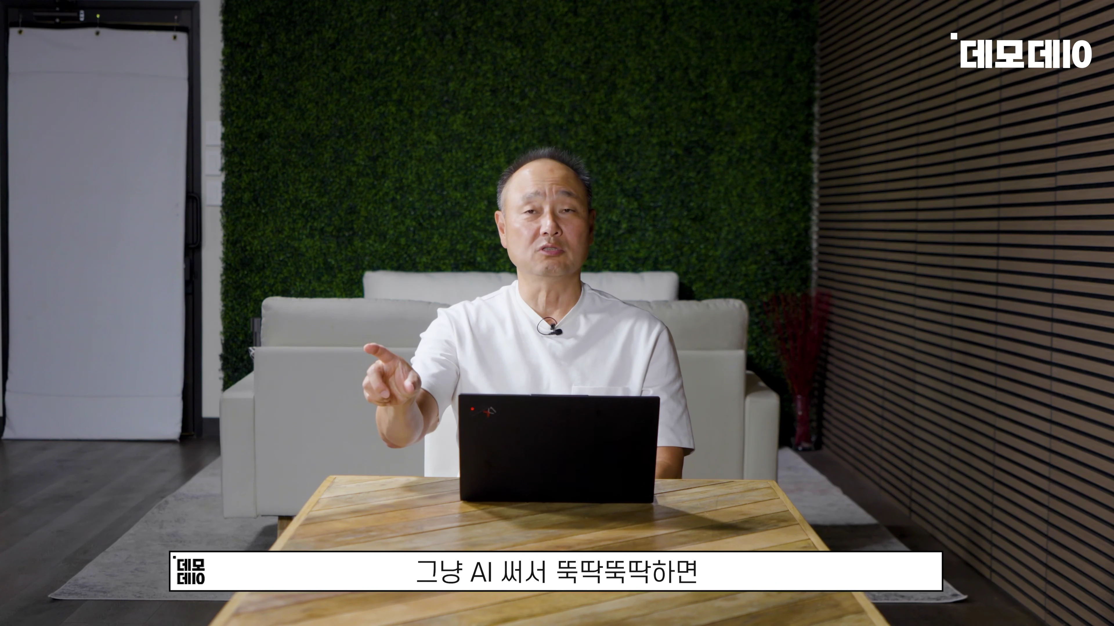 흰색 티셔츠를 입은 중년 남성이 책상 앞에 앉아 손가락으로 가리키며 이야기하는 모습
