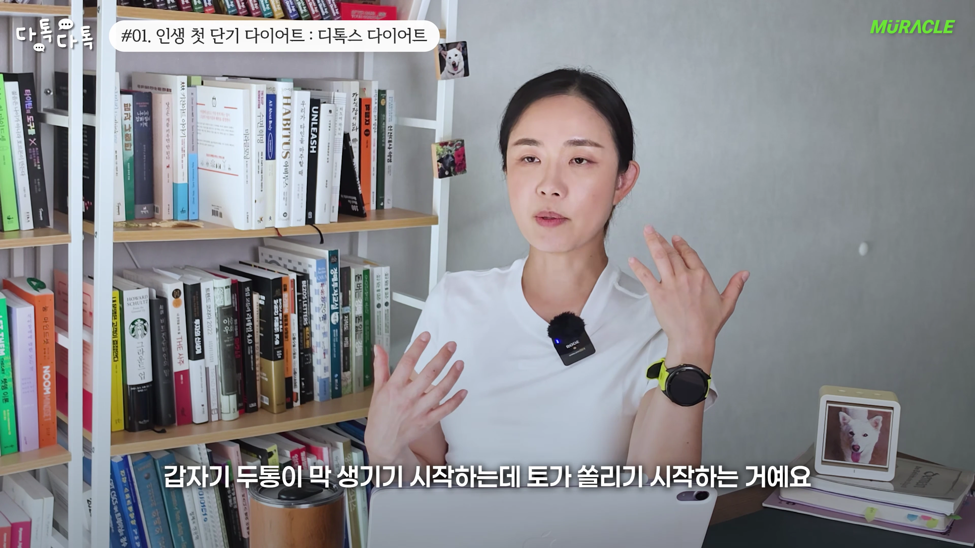 흰색 티셔츠를 입은 여성이 책장이 있는 실내에서 손짓하며 이야기하고 있다.