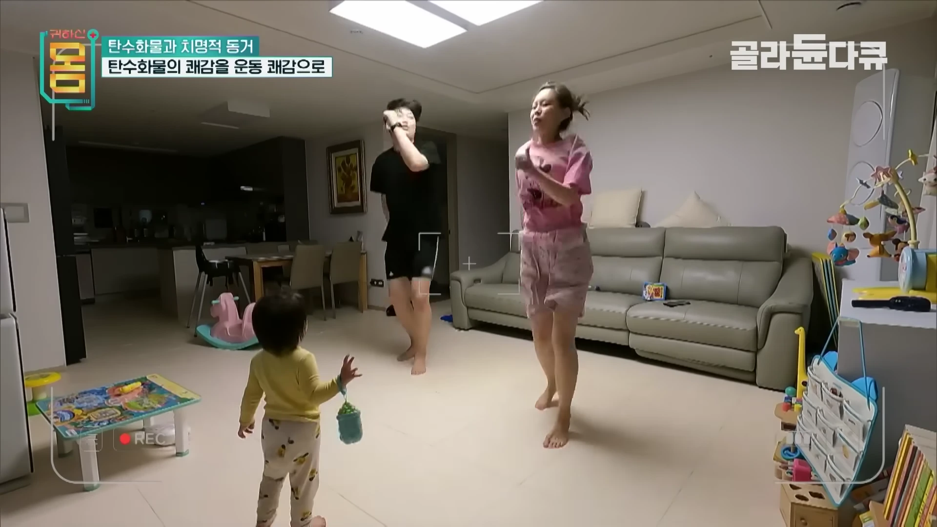 거실에서 편한 복장을 한 부부가 아이가 지켜보는 가운데 함께 제자리걸음 운동을 하고 있는 모습