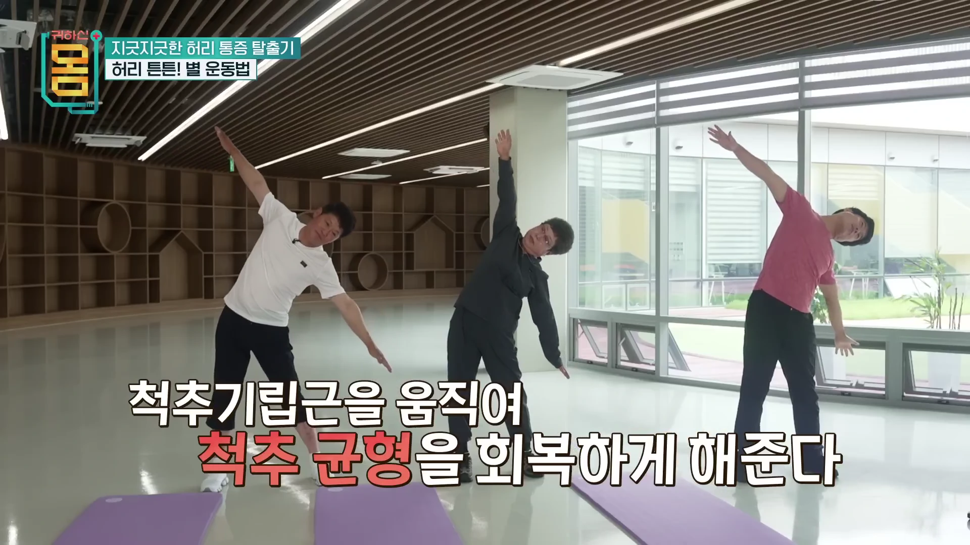 실내에서 세 사람이 팔과 다리를 넓게 벌리고 옆으로 몸을 기울이는 스트레칭 동작을 하고 있다.