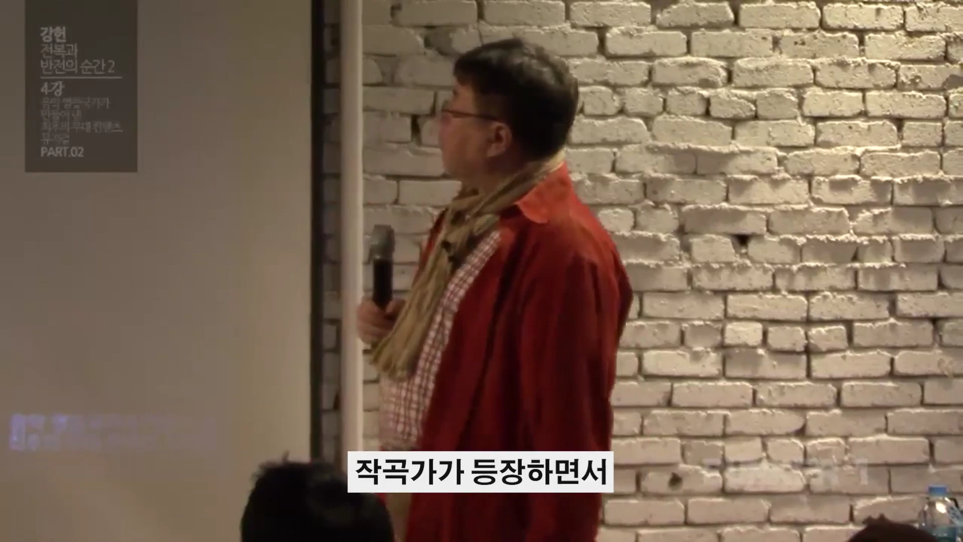붉은 셔츠를 입은 남성이 마이크를 들고 흰 벽돌 벽 앞에서 강연하는 모습