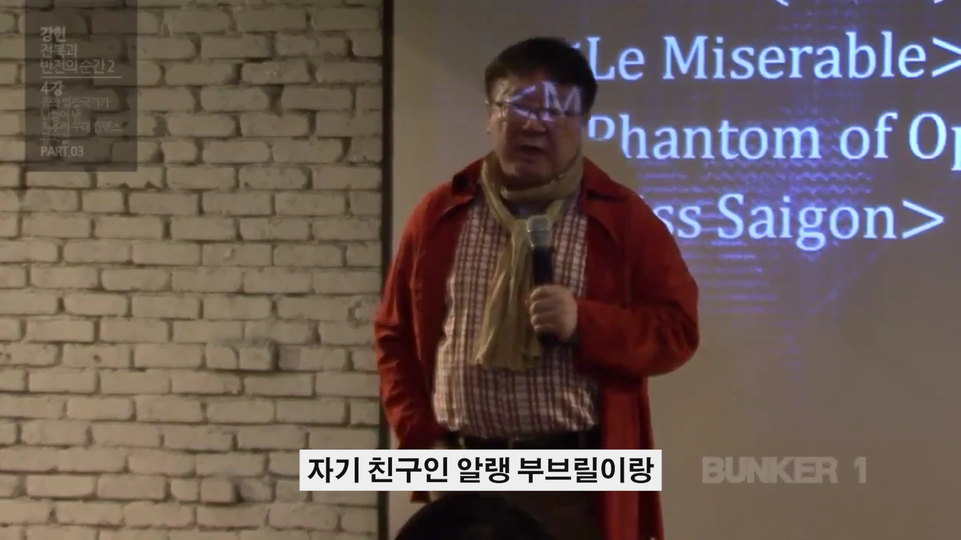 벽돌 벽을 배경으로 마이크를 들고 강연하는 남성과 뒤편의 스크린
