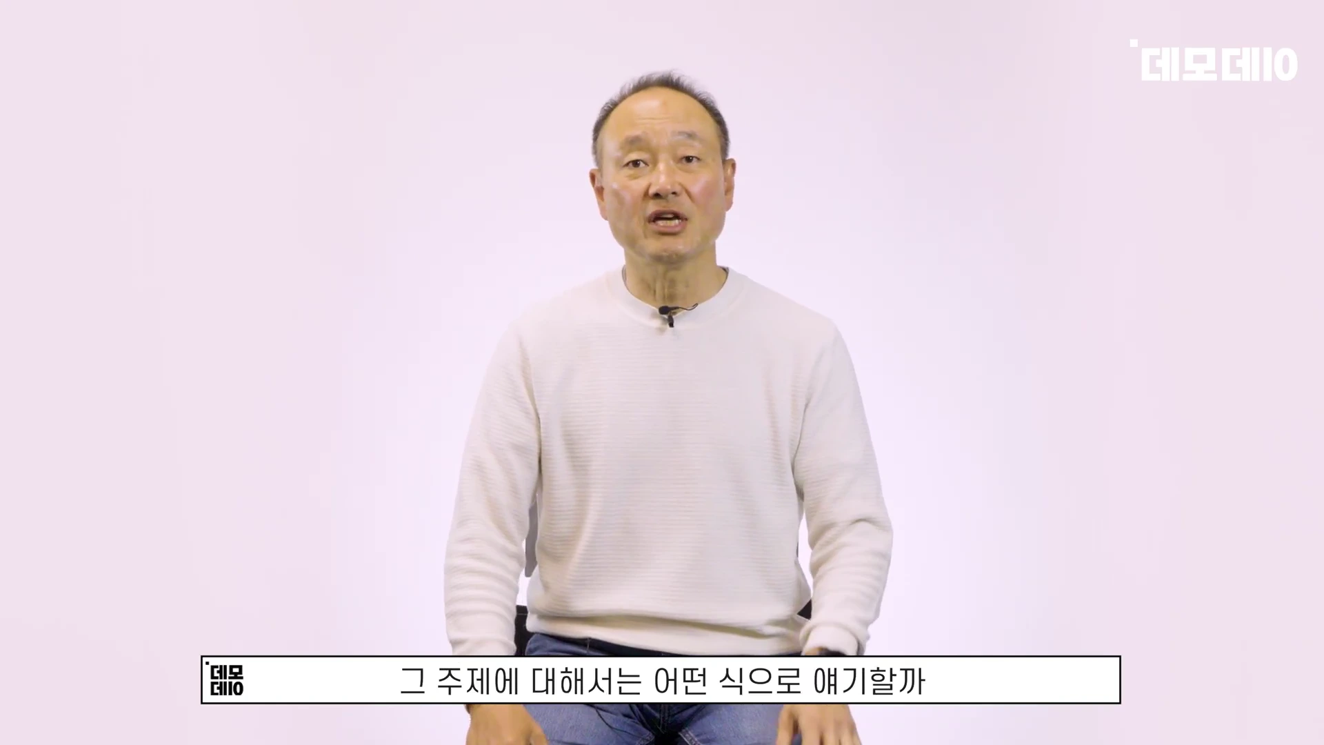 흰색 배경의 스튜디오에서 밝은색 니트를 입은 중년 남성이 카메라를 보며 이야기하고 있다.