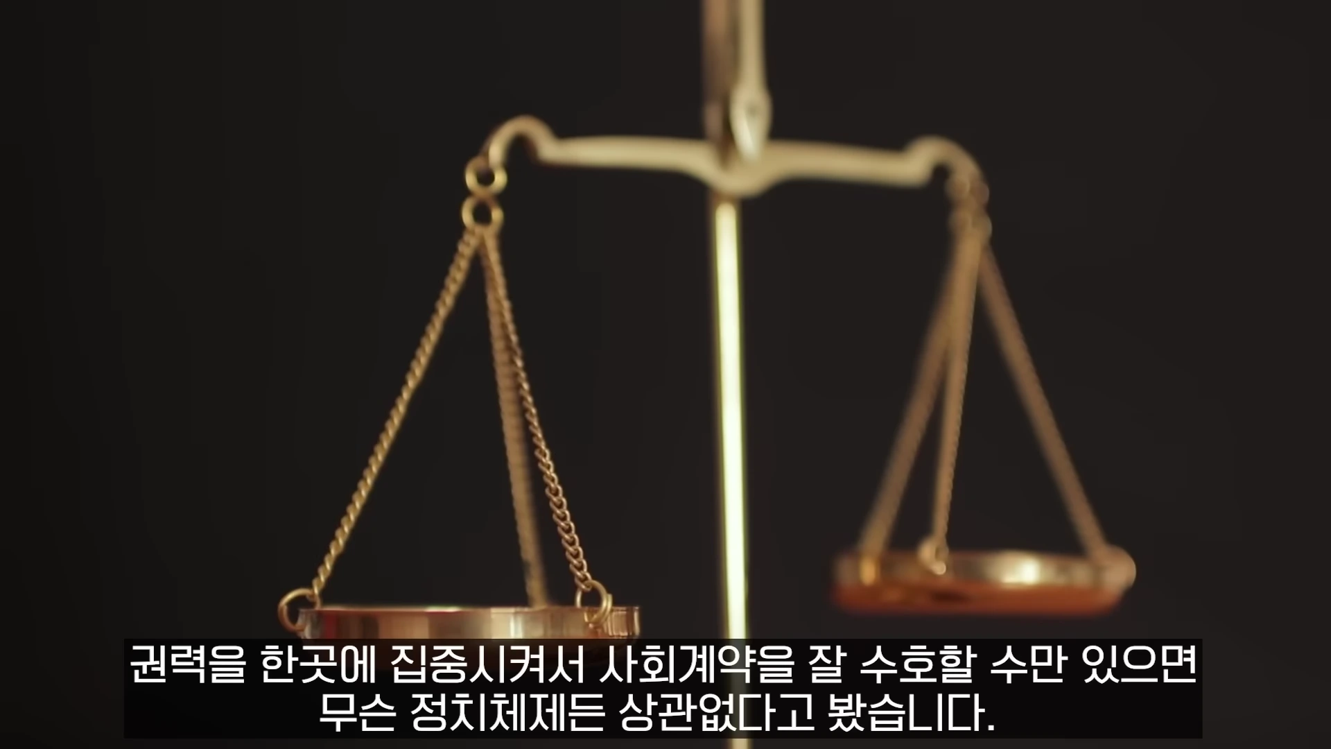 검은 배경 앞에 놓인 황금색 천칭 저울과 하단에 자막이 표시된 화면