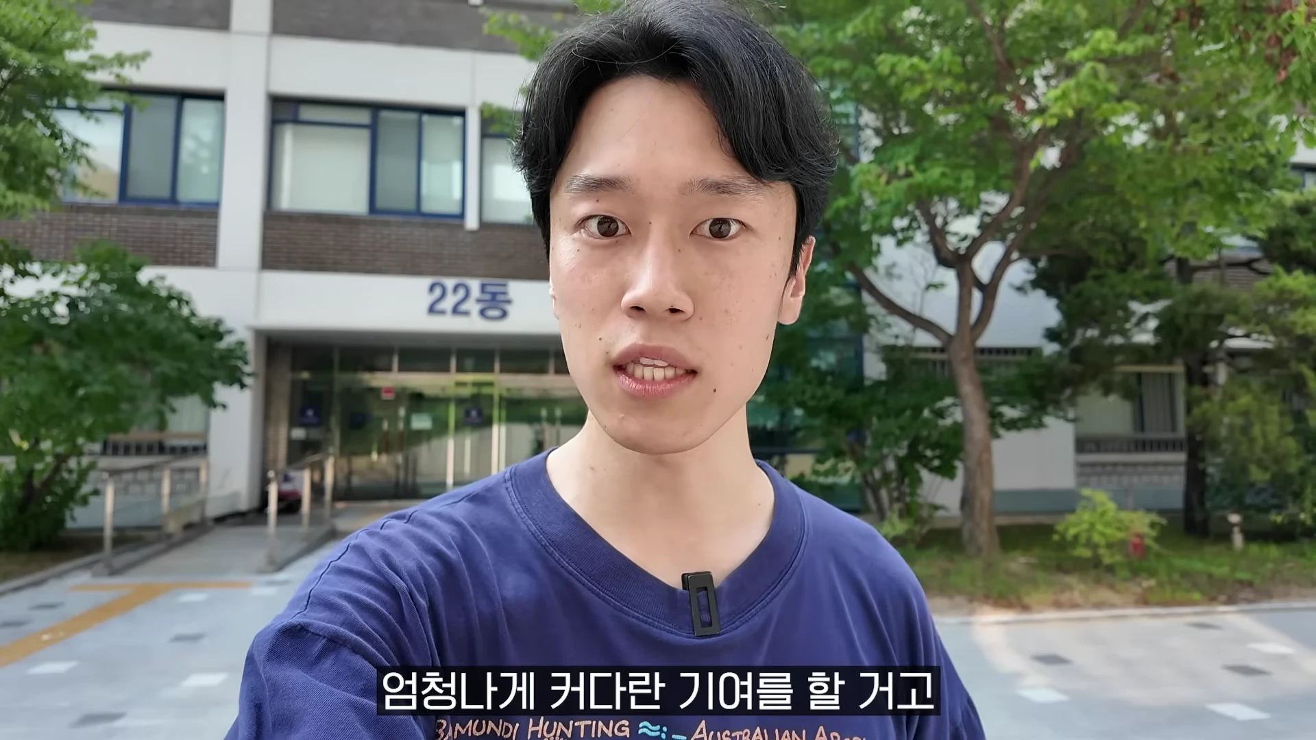 야외 건물 앞에서 카메라를 응시하며 이야기하는 남성