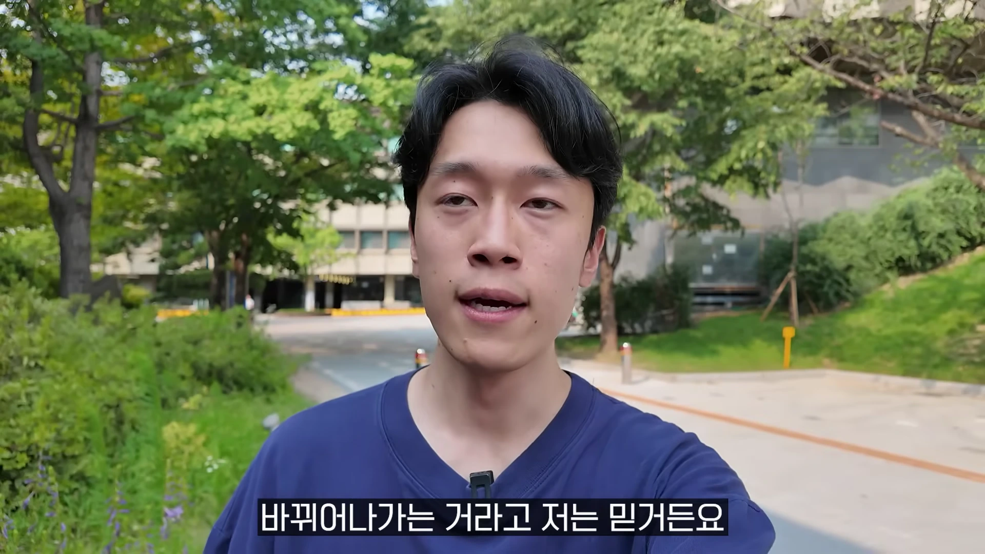 야외에서 남색 티셔츠를 입은 남성이 카메라를 보며 이야기하고 있다.