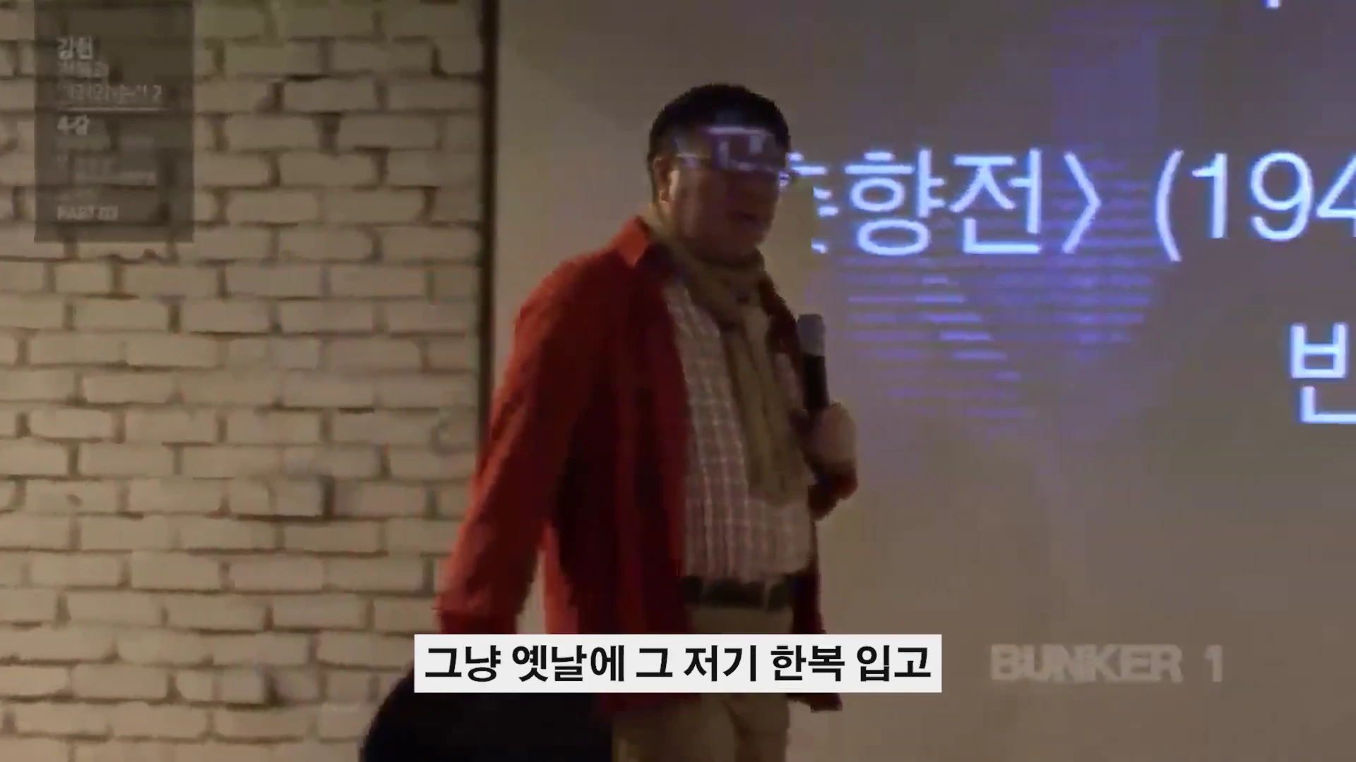 강연자가 마이크를 들고 벽돌 벽 앞의 스크린 옆에 서 있는 모습