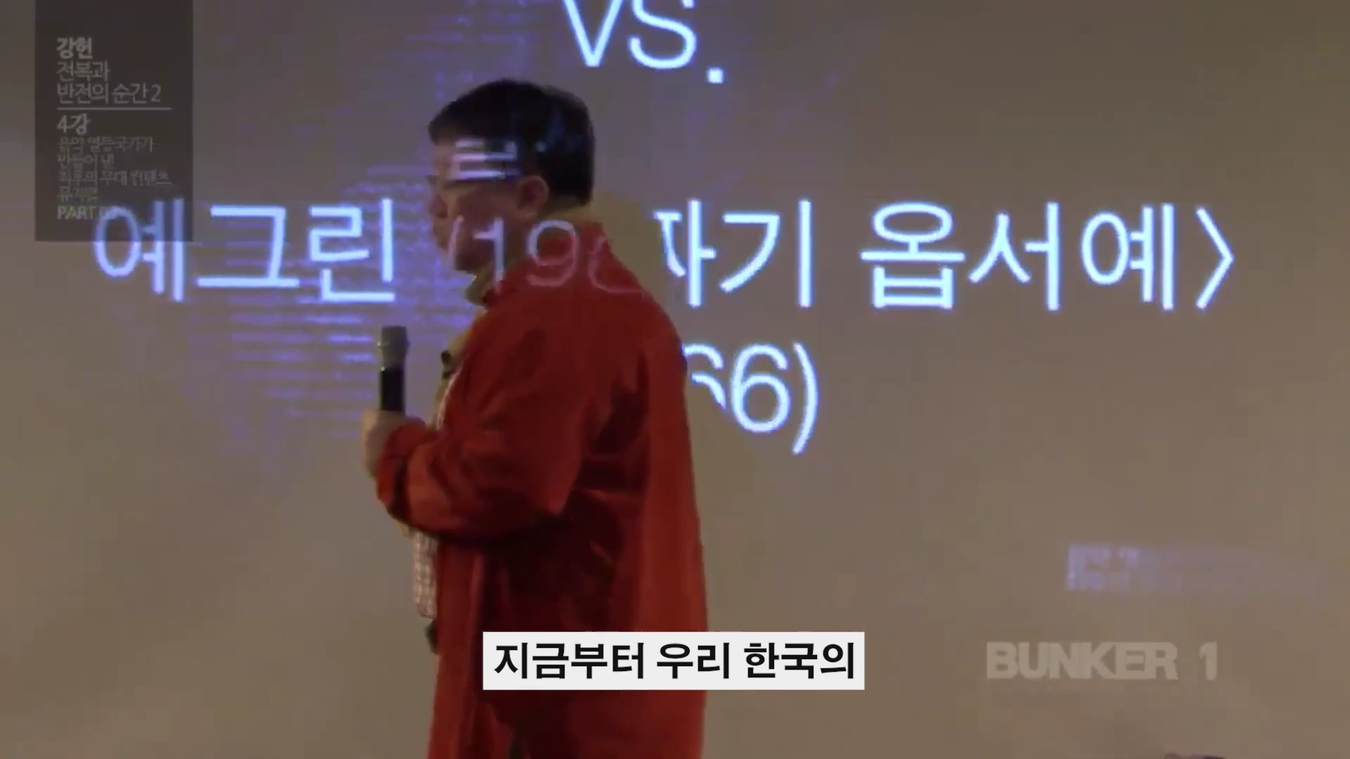 강연자가 마이크를 들고 스크린 앞에 서서 발표하는 모습